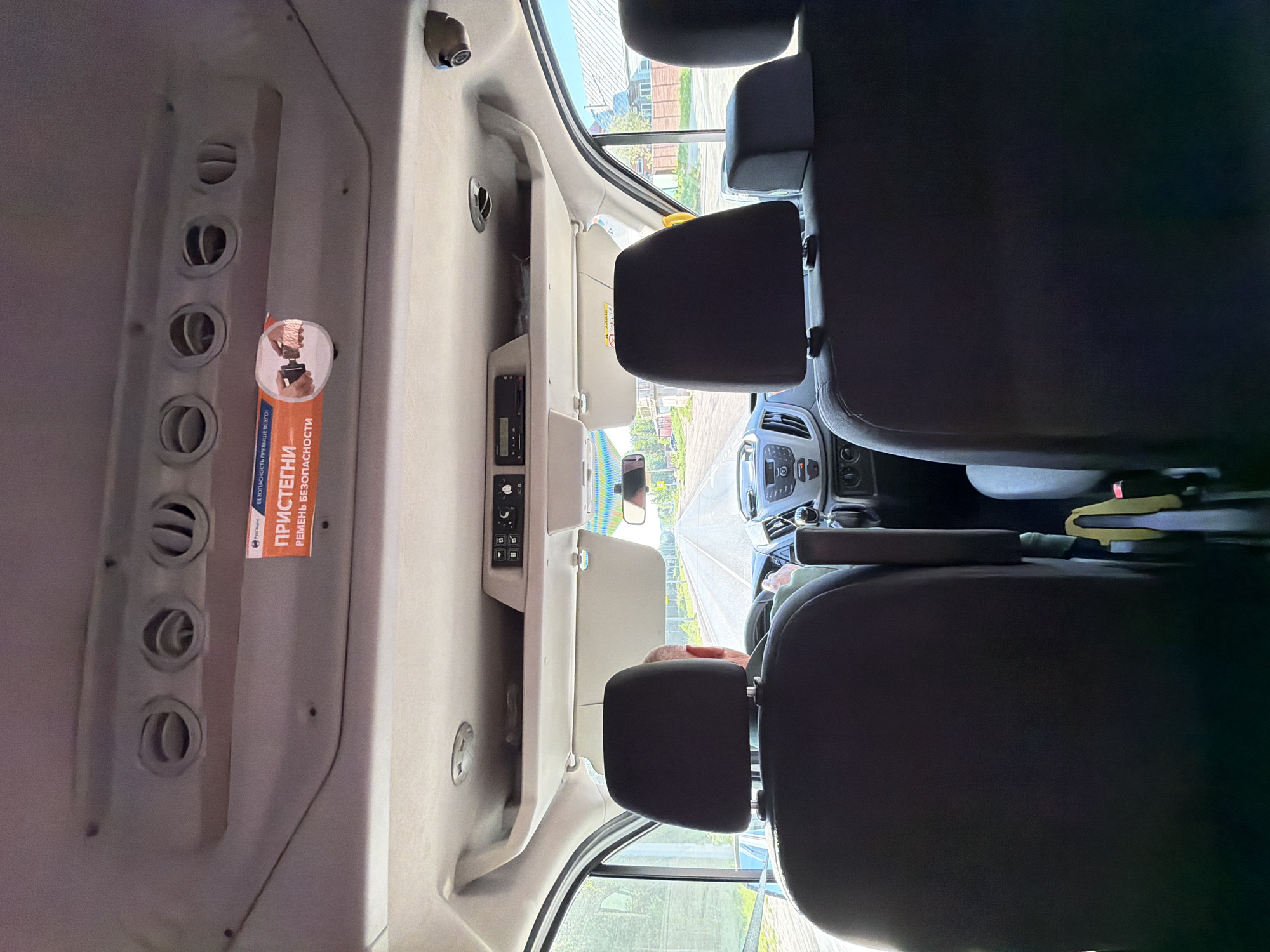The image shows the interior of a van or bus, possibly a passenger transport vehicle, likely in Chaikovsky, Russia, given the Russian text on the safety sticker. The perspective is from the rear, looking towards the front. There are passenger seats, a dashboard with controls, and air vents in the interior. The view through the windshield reveals a street. The presence of a driver is implied.