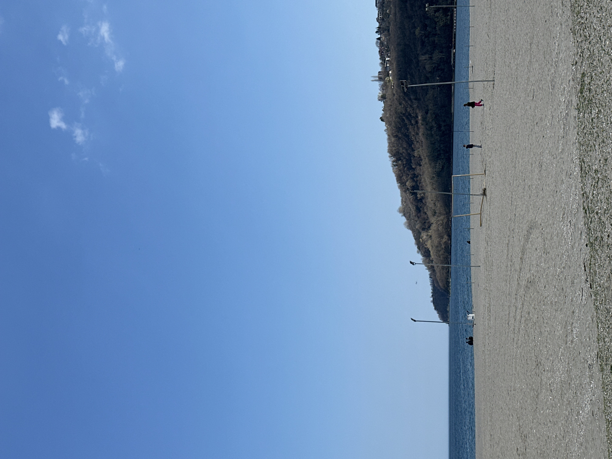 The scene depicts a beach along the coastline in Varna, Bulgaria. The sky is a clear, bright blue with a few scattered white clouds. A forested hill rises adjacent to the beach, topped with buildings. The beach itself is sandy with an area where it transitions to what appears to be small pebbles or shells. Lampposts are sparsely arranged along the beach near the water's edge. There are two people visible on the beach: one dressed in dark clothing, and another further away, dressed in pink and black. A set of soccer goal posts is located on the sand near the water.