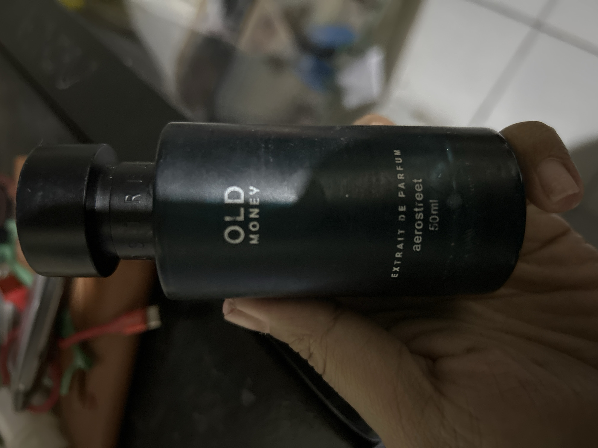 A close-up, eye-level, vertical shot shows a dark blue perfume bottle held by a person's hand in Depok, Indonesia. The bottle has a black cap and text on its surface that reads "OLD MONEY," "EXTRAIT DE PARFUM," "aerostreet," and "50ml." The background is blurred and appears to be an indoor setting with various objects.