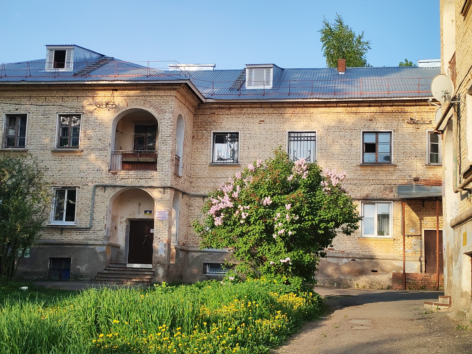The image shows a two-story brick building in Izhevsk, Russia. The building is painted yellow and has a blue metal roof with several dormer windows. A satellite dish is mounted on the right side of the building. The lower level has a partially submerged window. There are flowering lilac bushes in the front of the building and an area of grass and dandelions. A paved path leads towards the building.