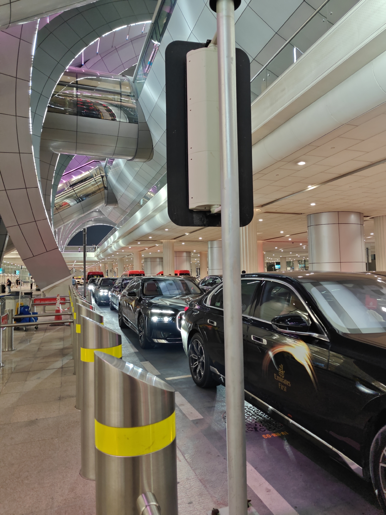 The image captures a scene at Dubai International Airport (DXB), specifically at the departures area. A line of black luxury sedans, identified by the "Emirates First" branding on the closest vehicle, are waiting. The airport's modern architecture is visible in the background, characterized by curved, metallic structures and prominent use of purple and white lighting. Red-topped vehicles, likely shuttle buses, are also present further down the line. People are sparsely visible in the distance, moving around the airport. The ground is paved, and metal bollards with yellow reflective bands line the waiting area. A traffic signal light is positioned in the foreground, partially obstructing the view.
