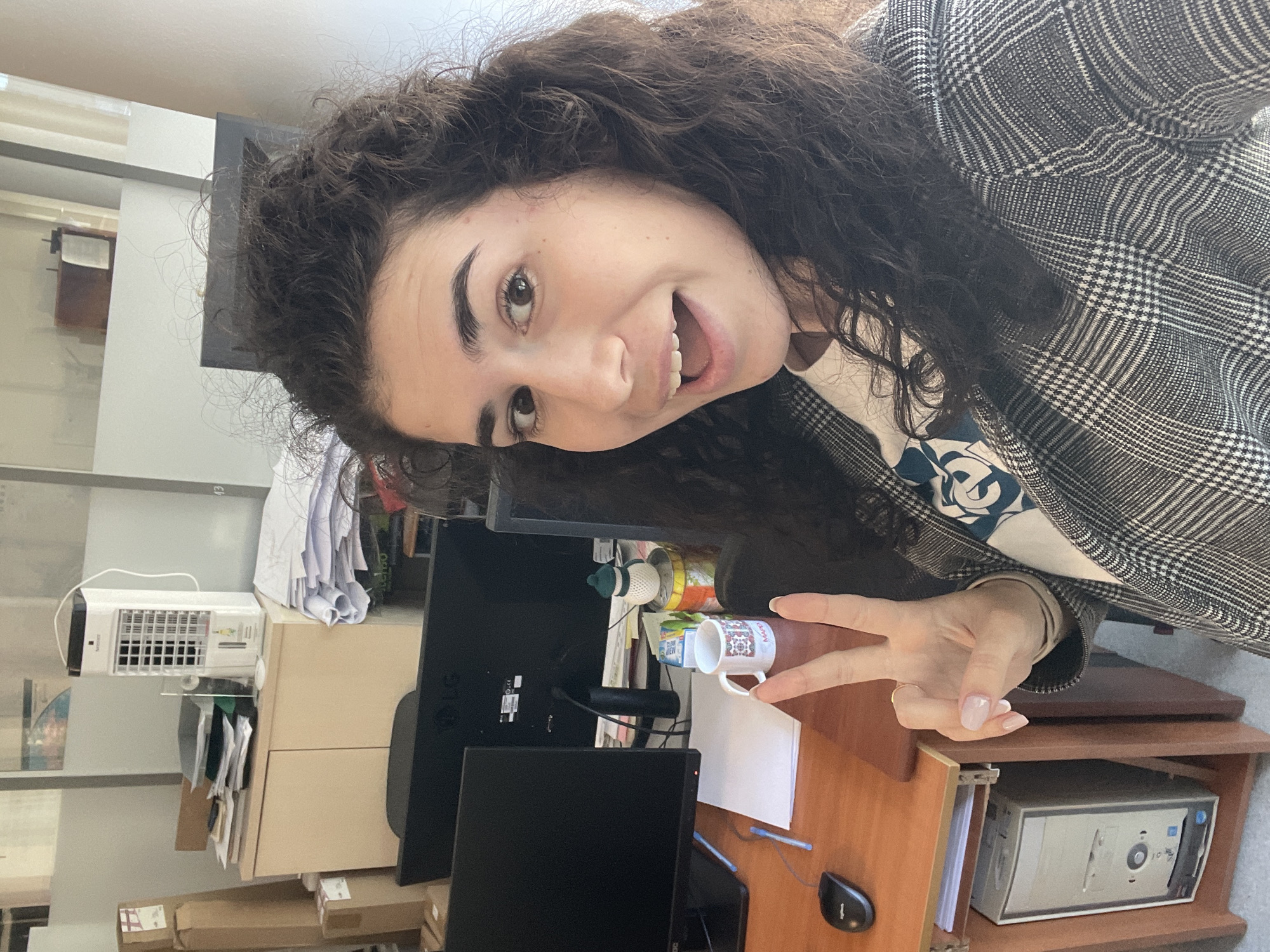 A woman with dark curly hair is smiling at the camera, holding up a peace sign with her right hand. She is wearing a gray and white plaid blazer over a white shirt. She appears to be indoors, in an office setting, as there is a desk with computer monitors, a mouse, pens, a mug, and a computer tower visible. There is also a filing cabinet with an air purifier on top of it. The location is identified as Sofia, Bulgaria.