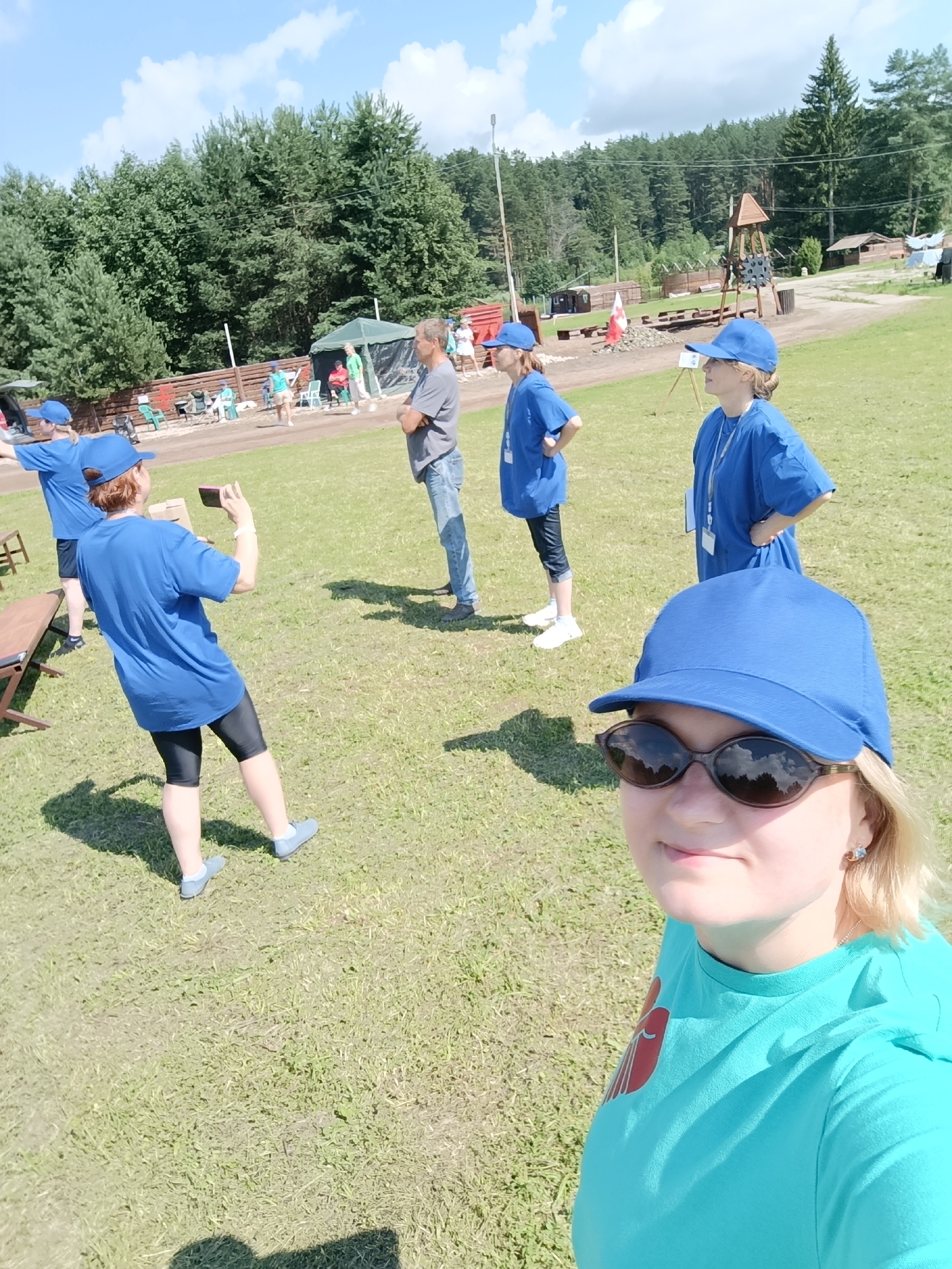 In a sunlit, grassy outdoor area, possibly a park or recreational facility, four individuals are visible, with a fifth person in the foreground taking a selfie. The setting is rural, with a dense forest bordering the area, and a wooden playground structure with a slide and other elements is visible in the background. Several tents and wooden structures are also present.

In the foreground, a young woman with blonde hair, wearing a bright blue t-shirt and a blue baseball cap, smiles at the camera, her face filling the lower portion of the frame. She is wearing dark sunglasses.

Behind her, three other individuals, two women and one man, are engaged in various activities. Two women, dressed in matching blue t-shirts and blue baseball caps, are standing and looking in different directions. One of them is holding a pink smartphone and appears to be taking a picture. The third individual, a man, is standing with his arms crossed, looking away from the camera. He is wearing a grey t-shirt and blue jeans.

The overall atmosphere is casual and appears to be a daytime outdoor gathering or event. The location is identified as being in the city of Prikamskii, Russia.