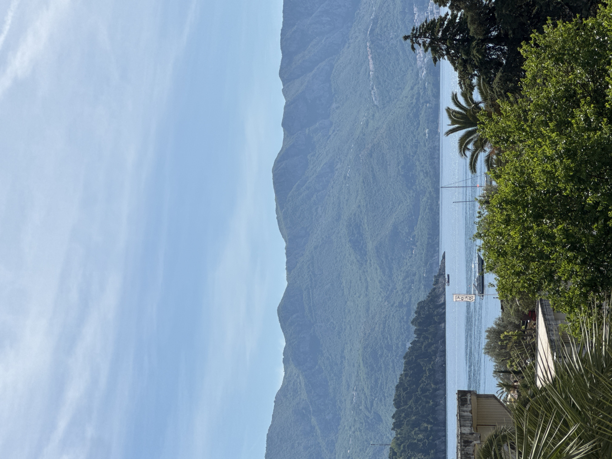 The image shows a view of the sea and mountains in Budva, Montenegro. The sky is light blue with streaks of clouds. The mountains are green and stretch along the coastline. In the foreground, there is vegetation and a building. A sign with the word "hulu" or "hoho" is visible. A boat is on the water.