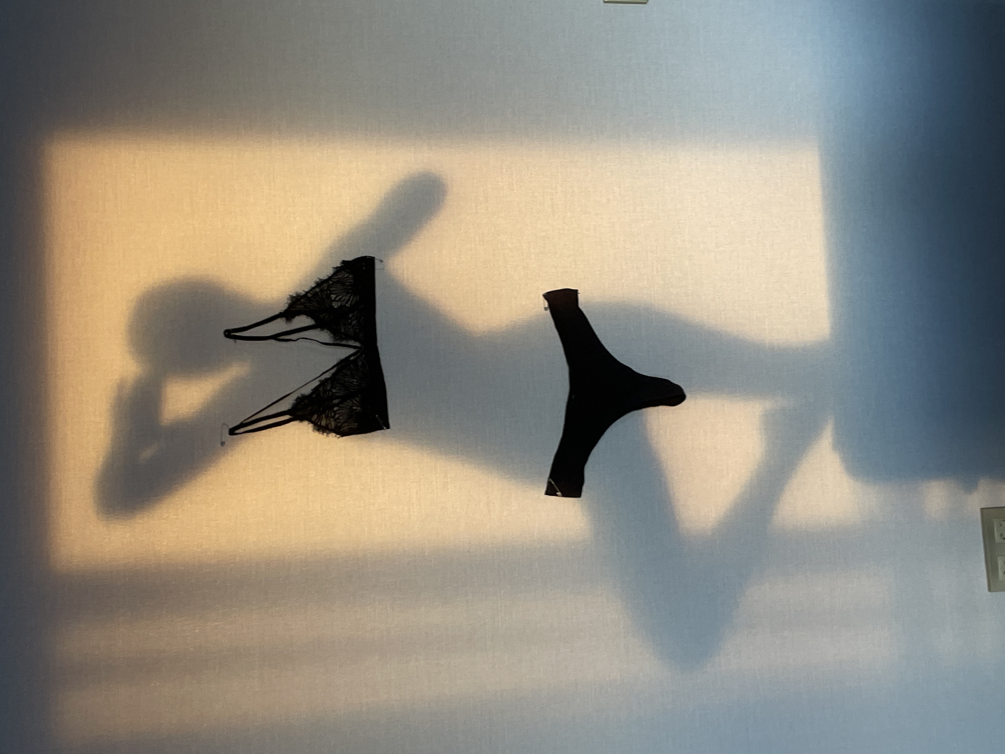 The image shows a white wall with shadows and two pieces of black lingerie hanging on it: a bra with lace detailing on the left and a thong on the right. The light source appears to be coming from the left, casting strong shadows. No people are visible. The background is a neutral-colored wall, with what looks like an electrical outlet or switch on the right side. There is no indication of the location being Подшивалово, Russia.
