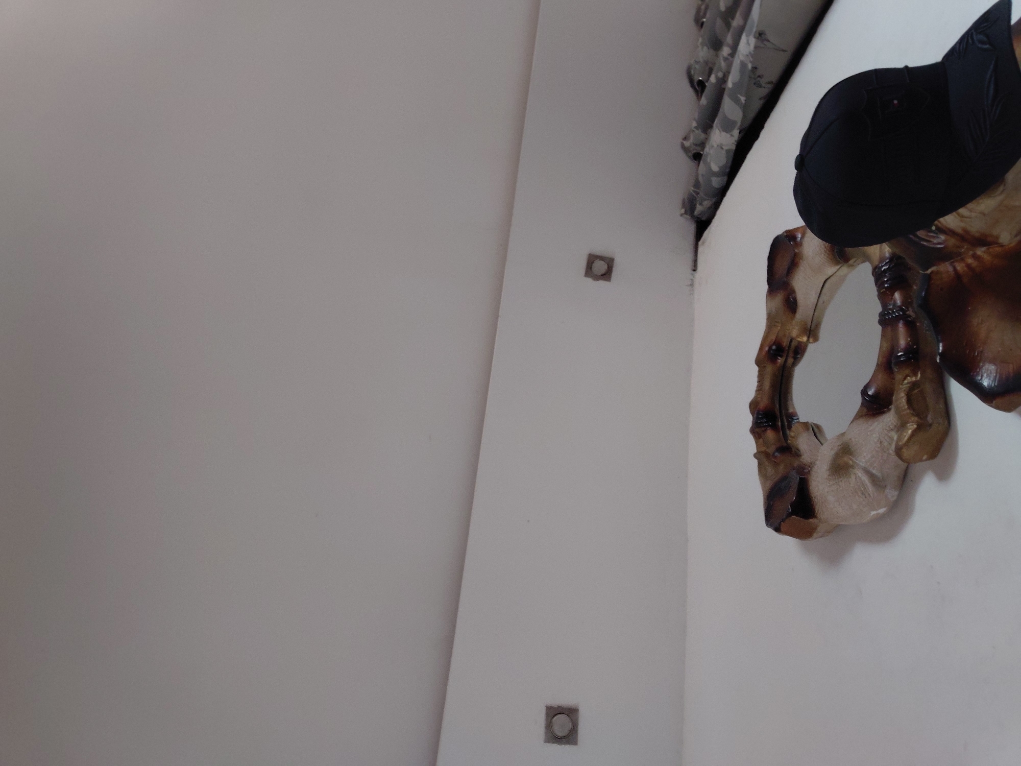 The image, appearing to be an indoor scene rotated 90 degrees clockwise, captures a brightly lit room with predominantly white walls. In the foreground on the right side, a unique, irregularly shaped wooden mirror frame with a circular mirror in its center is visible. The frame exhibits natural wood tones, ranging from dark brown to lighter beige, suggesting a carved or naturally formed piece. A black baseball cap is perched on top of this mirror frame.
Above the mirror and cap, a patterned fabric, possibly a curtain, hangs near the ceiling. The pattern on the fabric appears to be a grey and black camouflage or intricate design. Two square-shaped, metallic or silver-colored recessed fixtures, possibly lights or electrical covers, are embedded in the white wall; one is visible higher up, and another lower down towards the right. The walls are stark white, creating a clean and modern backdrop. There is no discernible activity taking place. The lighting is even and bright, suggesting either ample natural light or good artificial illumination. No text is visible in the image. The scene is located in Santa Ana, Venezuela.
