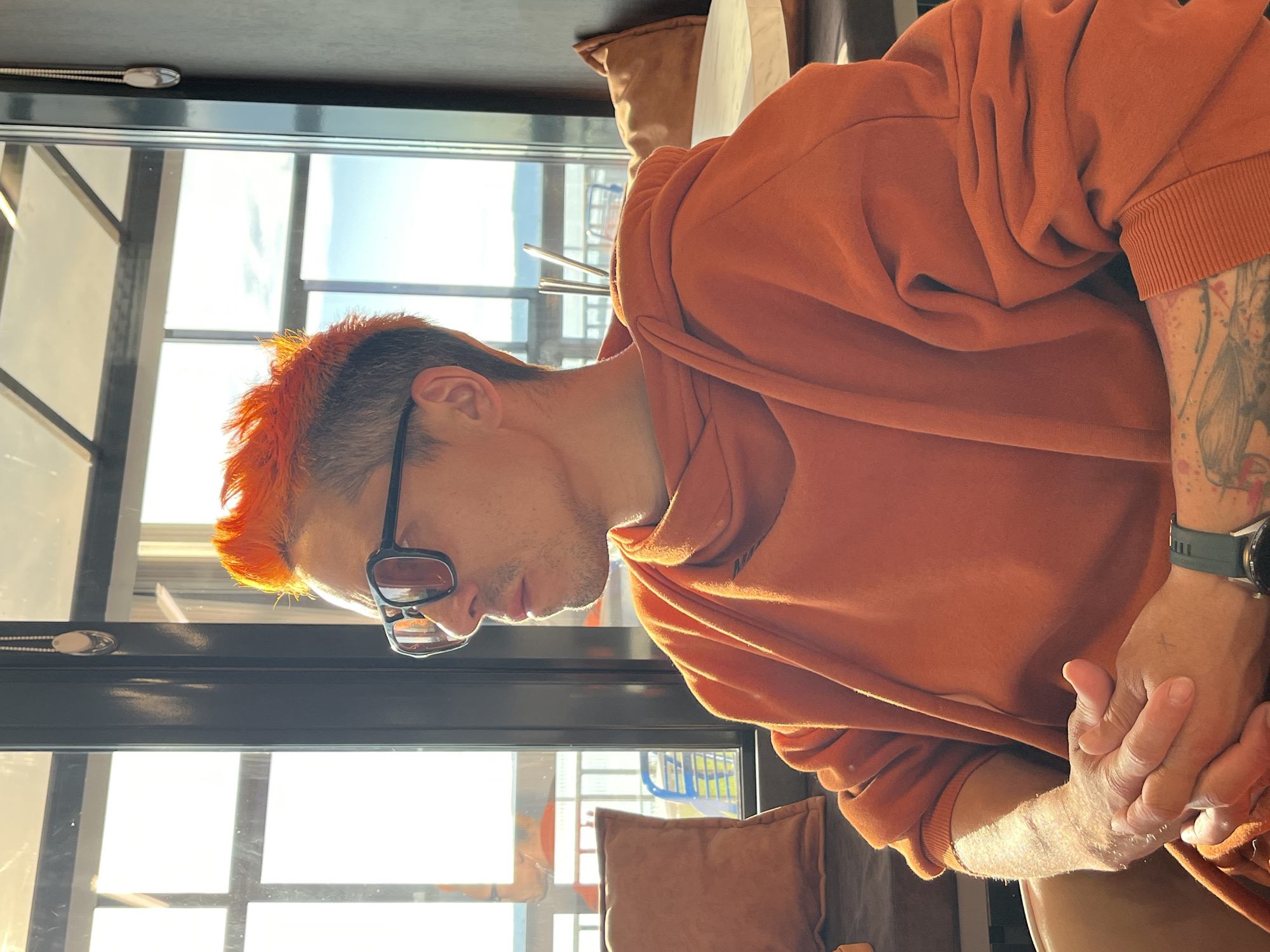 The image features a person, likely male, with distinctive bright orange, spiky hair. They are wearing a comfortable-looking orange hooded sweatshirt and dark-rimmed sunglasses with tinted lenses. The person is positioned in profile, looking downwards with their hands clasped or resting together in front of them, suggesting a moment of contemplation or quiet.
Visible on their right forearm and hand is an intricate tattoo, appearing to depict a mythical creature or winged design, with scattered red or pink splatters. A dark green watch with a black face is worn on their left wrist.
The setting appears to be indoors, likely a cafe, restaurant, or a room with large windows, given the strong natural light and the view of an outdoor scene. Bright sunlight streams in from the left, creating a luminous halo effect around the person's orange hair and highlighting the fabric of their sweatshirt. The backlighting casts the front of their face into shadow. Through the window, a bright, clear sky is visible, along with glimpses of distant structures or possibly a playground. An upholstered brown cushion is seen in the background.
The time of day is clearly daytime, characterized by the intense natural light, likely late morning or afternoon. No visible text is present in the image. The overall impression is one of a casual, reflective moment in a well-lit indoor environment in Chaikovsky, Russia.