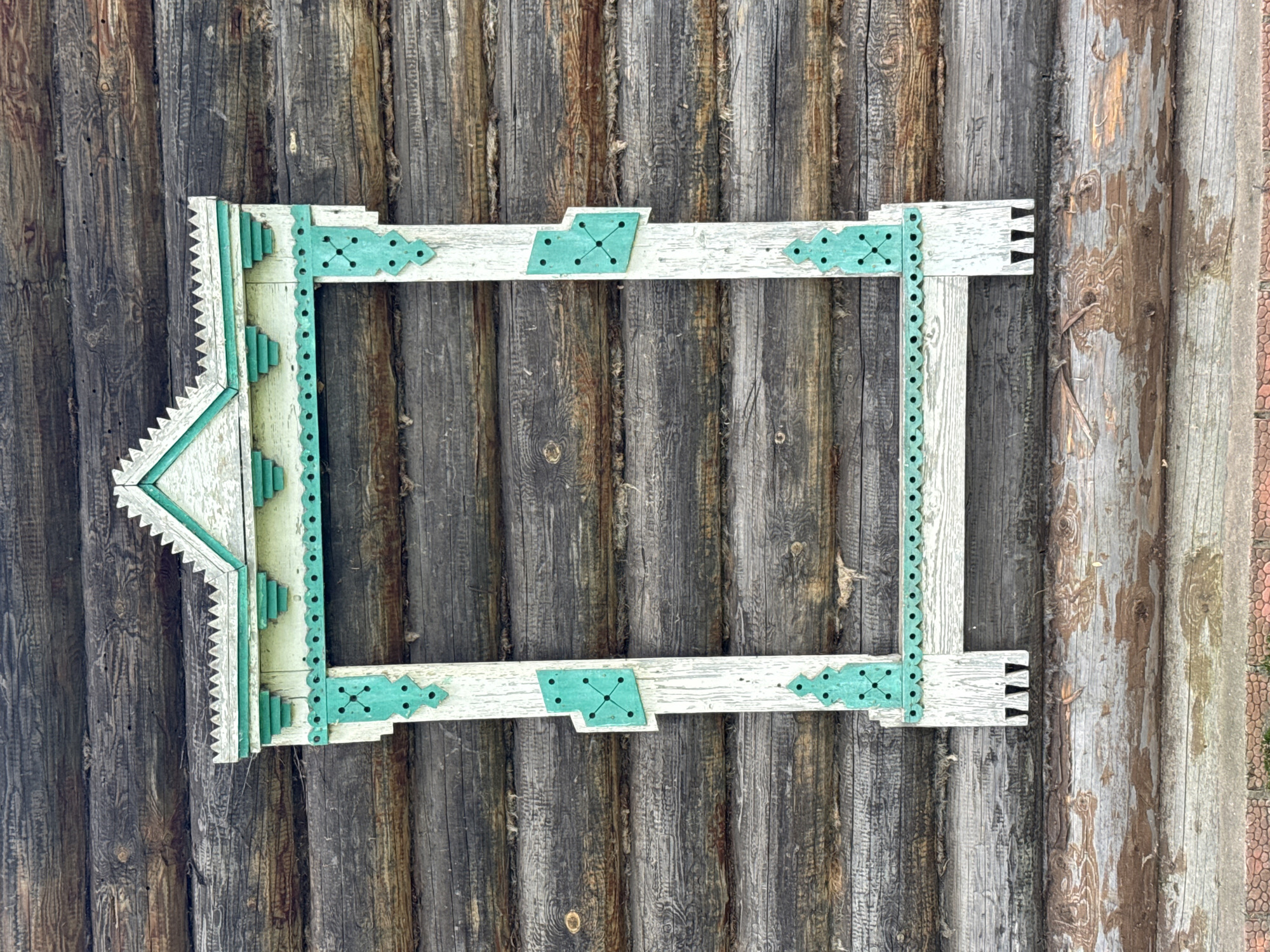 This is a vertical, medium shot of an ornate wooden window frame, painted white with teal accents, attached to the side of a log cabin. The log cabin's walls are constructed from horizontal, dark brown, weathered logs. The window frame has a decorative gable at the top with pointed, serrated edges, and vertical supports with teal decorative elements. The teal elements feature geometric patterns, including squares with four dots in the center. The bottom of the frame has two pointed, triangular cutouts. The background consists entirely of the log wall, with visible wood grain, knots, and weathering. The scene appears to be an outdoor setting, possibly in Votkinsk, Russia, given the architectural style which is characteristic of traditional Russian wooden houses. There are no people or actions depicted.