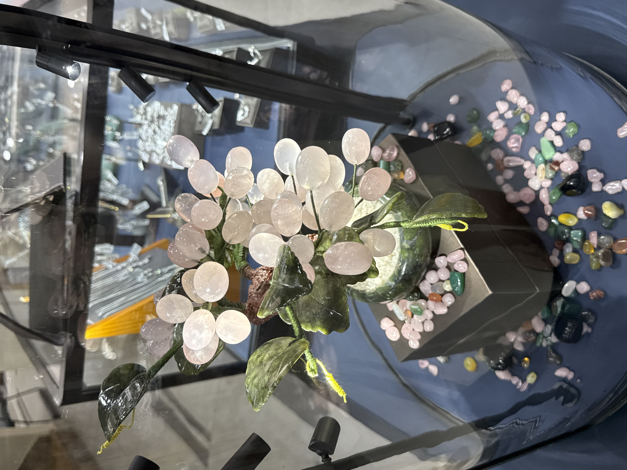 The image shows a decorative item, possibly a rose quartz tree, displayed inside a glass case in what appears to be a retail setting. The tree is composed of numerous small, round, pale pink stones resembling rose quartz, arranged to mimic a floral bouquet, with green leaves. The base of the tree is situated atop a dark, angular pedestal surrounded by a collection of small, multi-colored stones. The background reveals other jewelry displays and lighting fixtures, suggesting the location is a jewelry store or a similar retail environment. The lighting is bright, and the objects are clearly visible through the glass case. The location information provided indicates the scene is located in Nha Trang Ward, Vietnam.