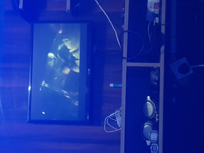The image shows a dimly lit interior with a blue hue. A monitor displays a blurry, dark image of a person holding a microphone. To the right, a shelf unit is visible with various objects including wires, headphones, and a small square device with a speaker. The overall impression is that of a cluttered, tech-heavy space.