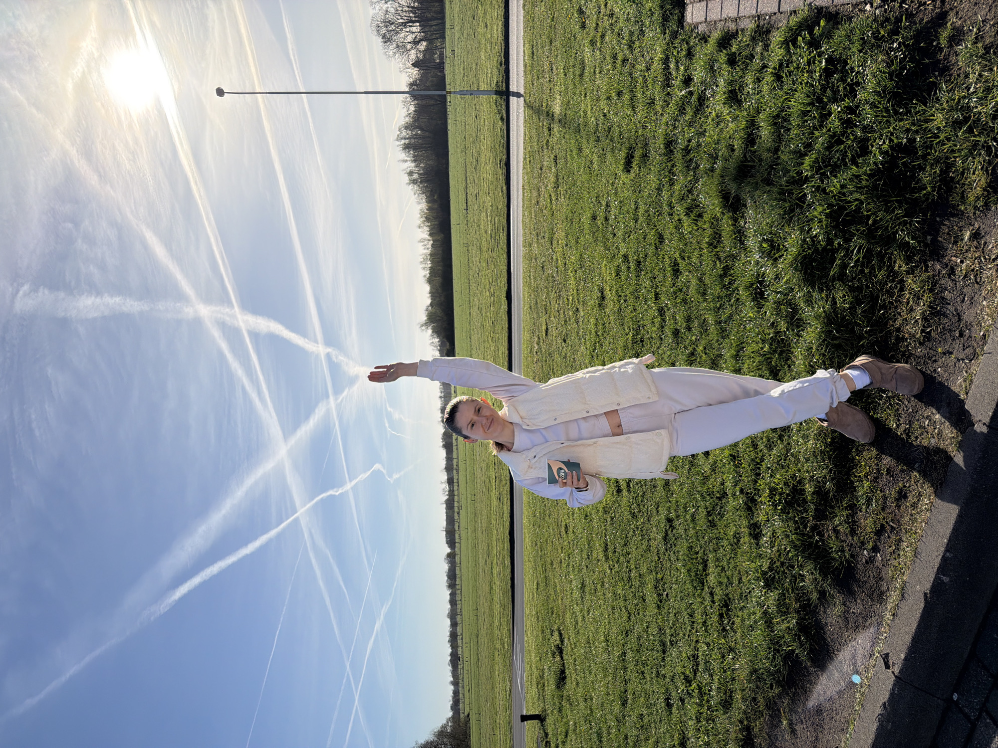 The image shows a young woman standing on a grassy field near a road in Klarenbeek, Netherlands. The woman is wearing a light-colored outfit, including a vest, pants, and shoes. She is holding a small, dark green rectangular object in her left hand and has her right arm extended upwards. Behind her is a green field with a line of trees in the distance. A street lamp is in the background as well. The sky is blue with numerous contrails. Part of the adjacent pavement and a brick edge are visible. The image is taken in daylight and seems to be rotated.