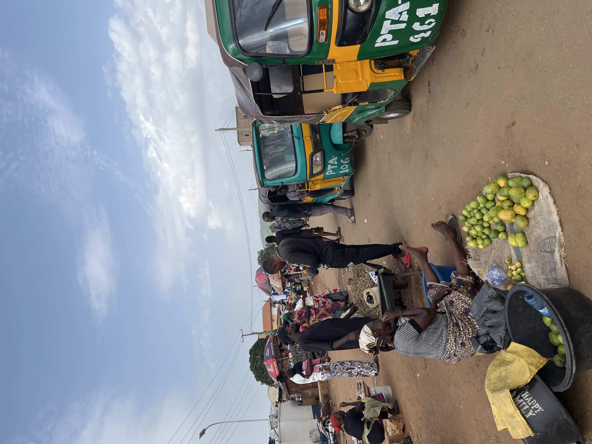This media file depicts a bustling street market scene in Jos, Nigeria. The primary subjects are a group of people, some appearing to be vendors and others customers, interacting within the market environment. In the foreground, a woman is seated, possibly resting or attending to a sale, with a pile of green fruits next to her. A large black basin filled with similar fruits and a bottle of water are also visible. Further in the background, several other individuals are seen standing or sitting amidst various market stalls and goods, with some of the stalls covered by colorful umbrellas.
Two distinct tuk-tuks, commonly known as "keke napep" in Nigeria, are parked side-by-side. Both are predominantly green and yellow, and one has the license plate "PTA 106" visible. The other tuk-tuk has "PTA 161" partially visible on its side. The overall setting is a dusty, unpaved street, characteristic of many Nigerian markets. The weather appears to be partly cloudy with a bright sky, suggesting it is likely daytime, possibly late morning or early afternoon. The mood seems to be one of everyday commerce and community activity.
Visible text includes the license plate numbers on the tuk-tuks: "PTA 106" and "PTA 161". There is also text on a yellow cloth that reads "HAPPY FAMILY".