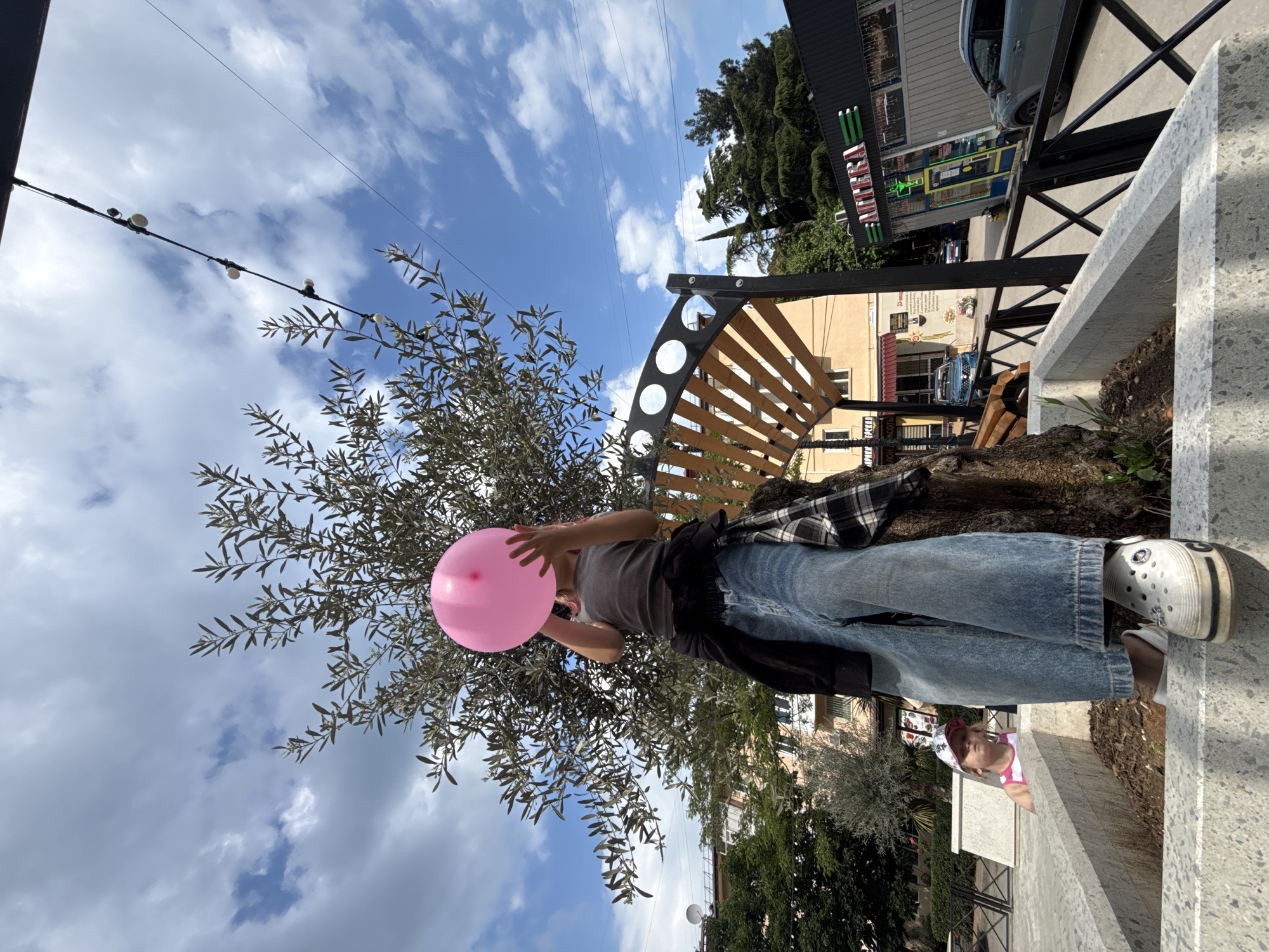 A low-angle outdoor shot captures a person, partially obscured by an olive tree, holding a pink balloon against a bright blue sky with scattered clouds. The person wears a gray tank top, baggy denim jeans, and white clogs. A black jacket is tied around their waist, and a plaid shirt is also tied around their waist. The person stands on a stone planter, looking up at the balloon. In the background, a young child wearing a pink and white cap and tank top peeks over a stone wall. Further in the background, buildings, trees, a black metal fence, and a pharmacy with a green awning are visible. String lights are hung above. The scene appears to be a public outdoor space.