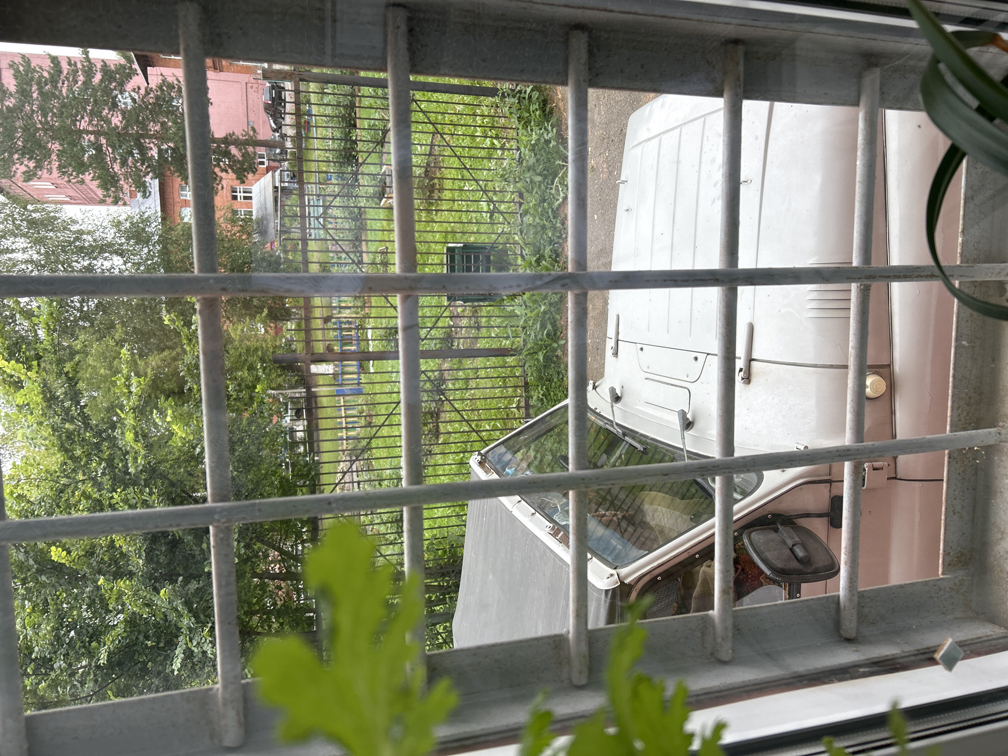 The image shows a view through a barred window, looking out onto an outdoor area. In the foreground, the top portion of a light-colored, somewhat dirty vehicle, possibly a UAZ-469, is visible. The vehicle has a soft top, and its windshield is open. A side mirror is attached to the driver's side door. Behind the vehicle, a metal fence encloses a grassy area with some scattered plants. Beyond the fence, there are trees and a tall, pink building with multiple windows, indicative of an apartment block. Further in the distance, another building with a red brick facade and white window frames is visible. The overall environment suggests a residential area. The presence of the barred window implies the view is from an upper floor or a secure building. The scene is set during daylight hours, with overcast sky conditions. The specific location is identified as Tomsk, Russia, which is consistent with the architectural style of the buildings and the climate suggested by the vegetation. There are no people clearly visible in the image.