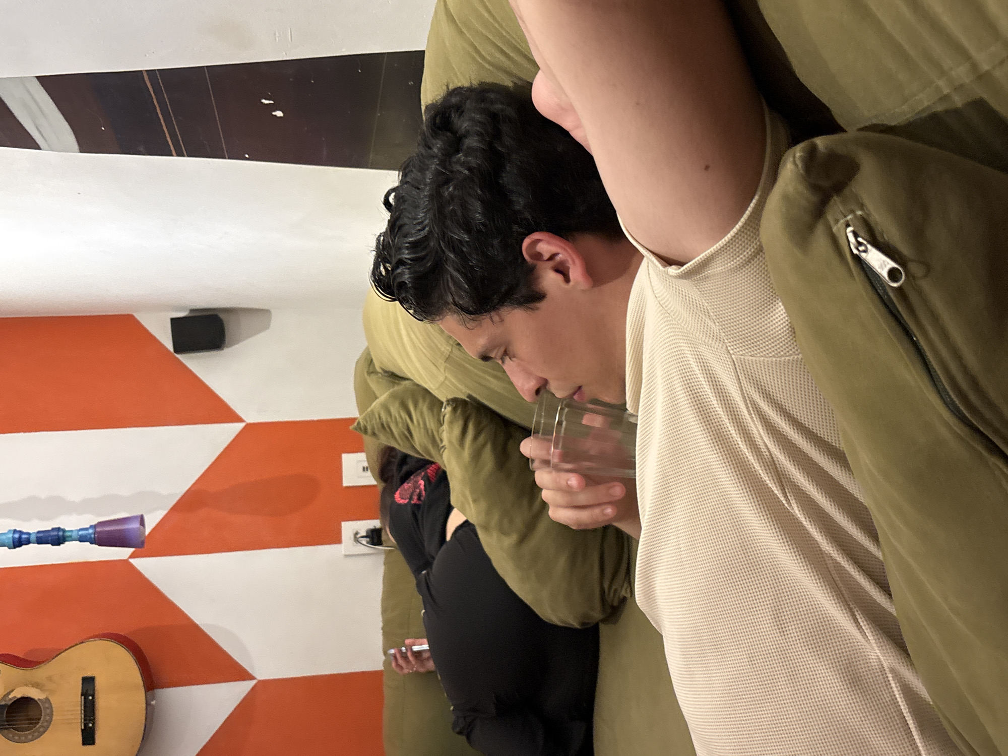 The image captures a scene indoors, potentially in a residential setting. A person with dark, curly hair is reclined on a large, olive-green couch and is holding a clear glass to their lips, possibly drinking. The person is wearing a light beige waffle-textured shirt and is partially covered by an olive-green jacket with a visible silver zipper. Another person, dressed in black, is partially visible lying on the couch behind the first person. The backdrop shows a white and orange patterned wall, with a wooden guitar hanging on it. A blue and purple object, possibly a decoration or an art piece, is also mounted on the wall. There is a black speaker affixed to the wall near the ceiling. The image is not clear enough to determine if the location is São Paulo, Brazil.