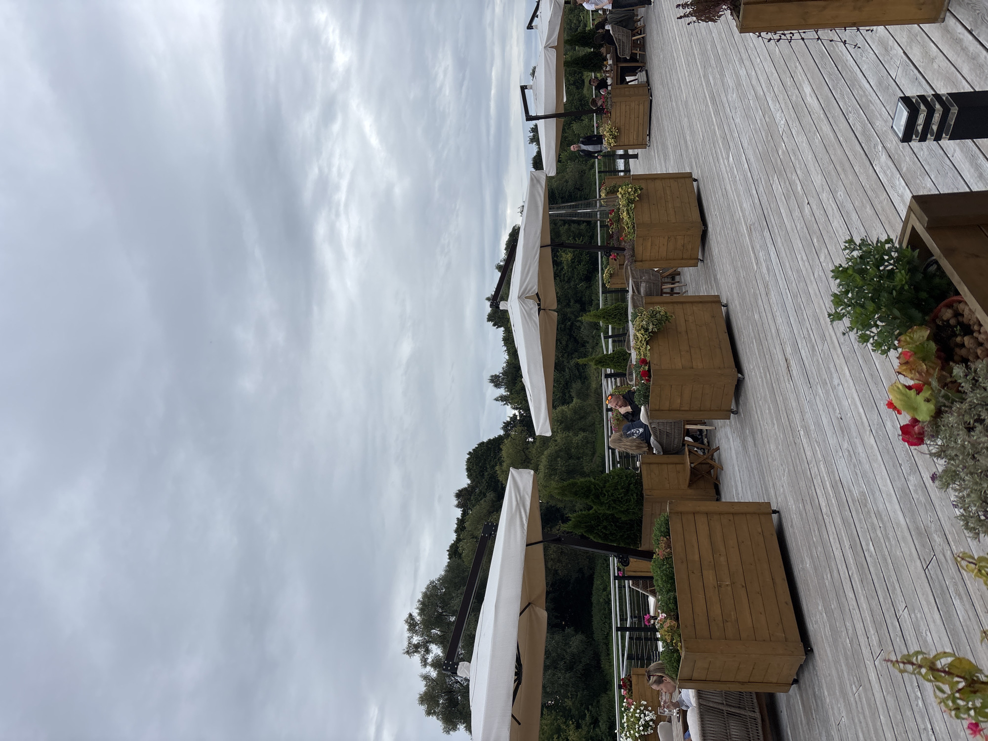 The image depicts an outdoor seating area of a restaurant or cafe, possibly located in Zvenigorod, Russia, with a view of a natural landscape. The scene is set on a wooden deck furnished with large white umbrellas providing shade over several tables and chairs. Wooden planters filled with greenery and flowers are strategically placed throughout the area, some containing small coniferous trees. Several patrons are seated at tables, engaged in conversation or dining. In the background, a dense line of trees borders a body of water, suggesting a scenic location. The sky is overcast, with a cloudy, muted grey hue. The overall atmosphere appears relaxed and serene.