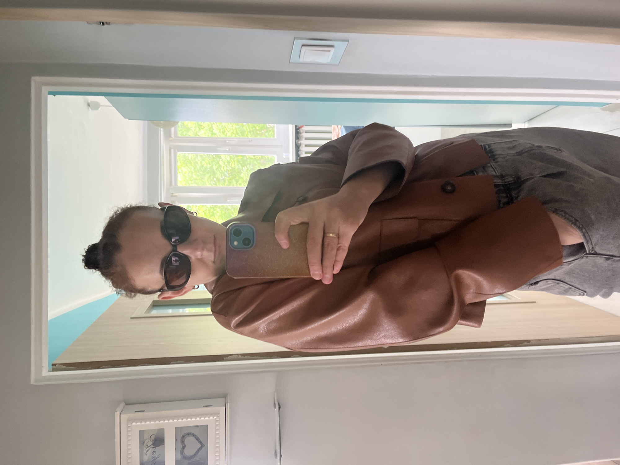 A person is visible in a mirror selfie, wearing sunglasses and a brown jacket over what appears to be a denim garment. The background shows an interior space with a window through which greenery is visible. A white framed object adorns the wall, while a light fixture is set into the ceiling. The location is unknown.