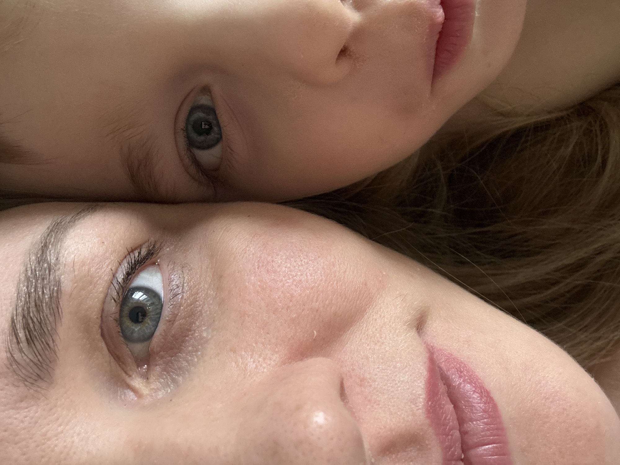 The image shows a close-up of two faces, positioned head-to-head. The upper face is that of a child, with fair skin, light hair, and a blue eye visible. The lower face appears to be an adult, with similar fair skin, and a blue eye also visible. Both faces are in focus, and the photograph appears to be taken in a neutral indoor lighting environment. The location cannot be identified.
