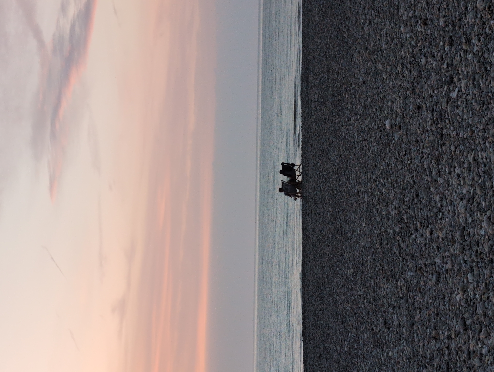Two individuals are seated in beach chairs on a pebble-covered shore, facing the sea. The sky above is a gradient of soft pink and pale orange hues, suggesting either sunrise or sunset. The sea is calm and appears as a muted blue-gray. The location is the beach in Mers-les-Bains, France, known for its colorful clifftop houses, though these are not visible in this particular frame. The scene conveys a sense of quietude and shared presence.