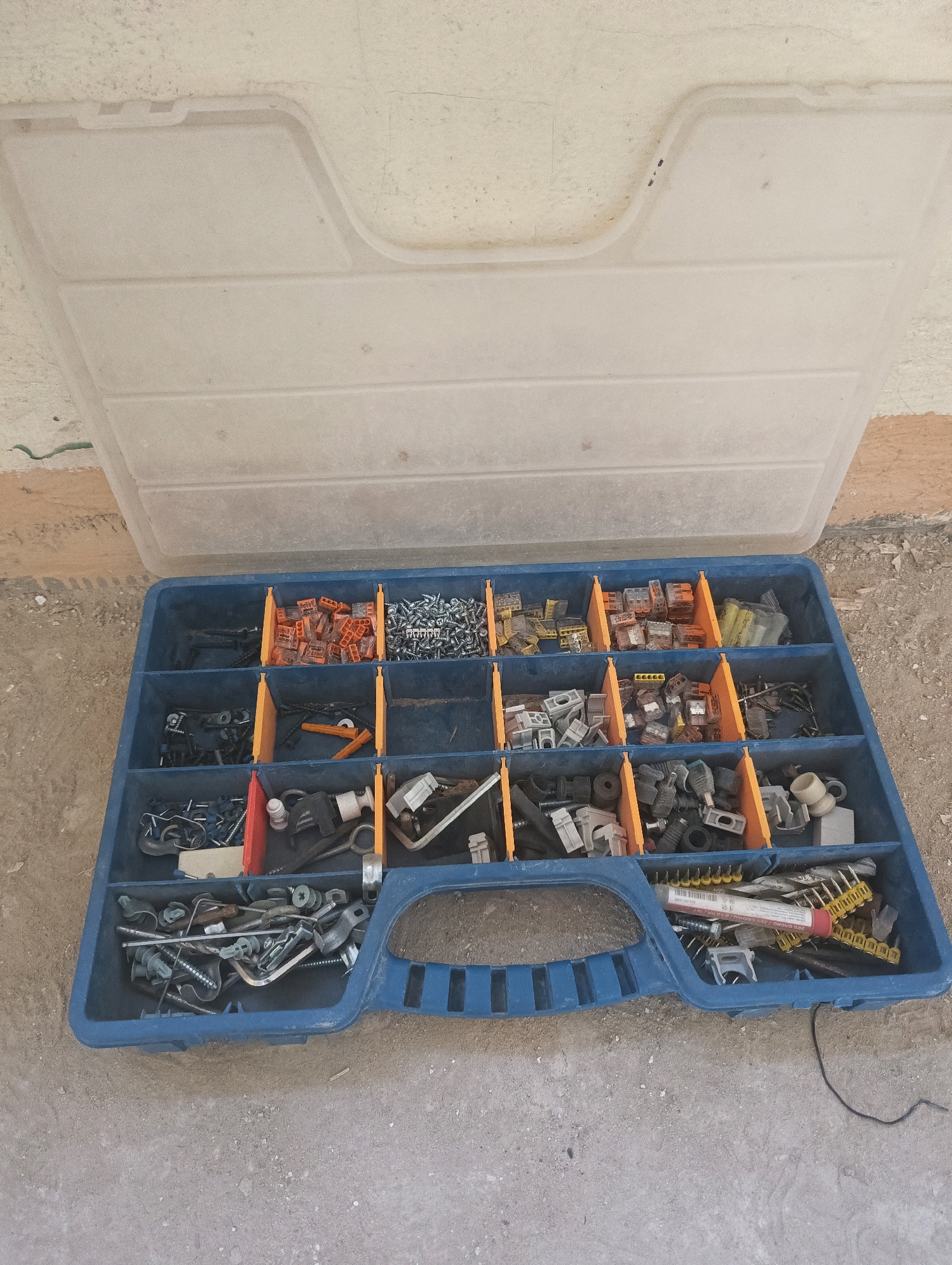The image shows an open, blue plastic organizer box filled with various hardware items and electrical connectors. The box is sitting on a concrete or dirt surface, likely outdoors, with a textured wall in the background. The lid of the organizer is open and tilted upwards, revealing multiple compartments containing an assortment of screws, wire connectors (some orange, some clear), small metal fittings, and what appear to be electrical components. The overall impression is of a functional storage solution for tools and supplies. The location is not discernible from the image itself.