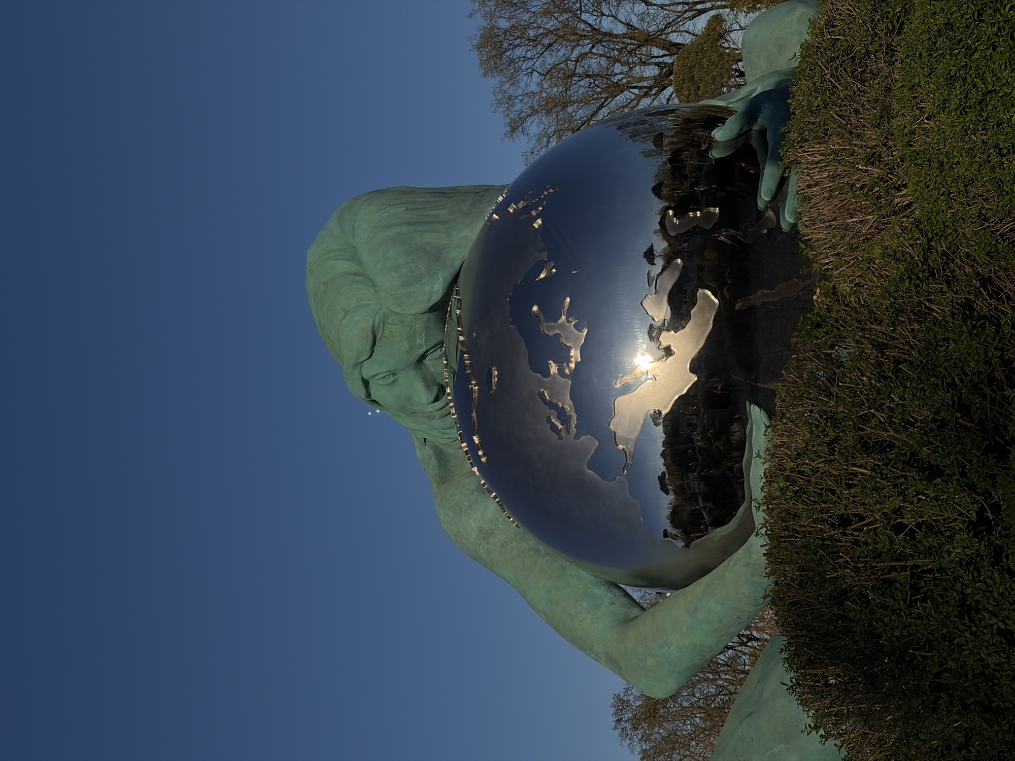 A low-angle, outdoor shot captures a green-patinaed bronze statue of a woman supporting a spherical, polished metal sculpture resembling the Earth. The statue is partially obscured by a thick, green bush on the right. The polished sphere reflects the environment, including the sun and sky. The sky is a clear, deep blue. Bare tree branches are visible in the background, indicating it is possibly late autumn or winter. The statue is located in the city of Krasnodar, Russia.