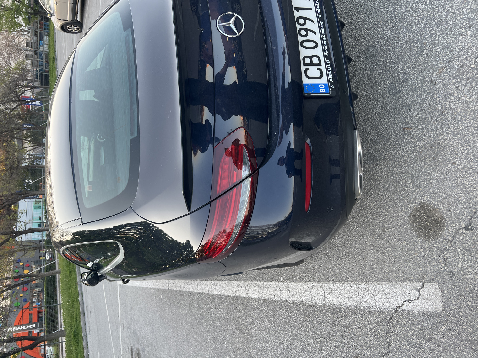 The image depicts the rear of a dark-colored Mercedes-Benz parked in a paved parking area. The car's license plate reads "CB 0991 BG". The make of the plate is ARNOLD Parsberg-Lupburg. White parking lines are visible on the pavement. The reflection on the car's surface suggests an urban environment. Visible in the background of the reflection are trees and what appears to be a children's play area and a climbing wall branded as 'DOMINOS'. Based on this information, it can be deduced that the location is likely in the city of Plovdiv, Bulgaria, near a park or recreational area. There are no people clearly visible.