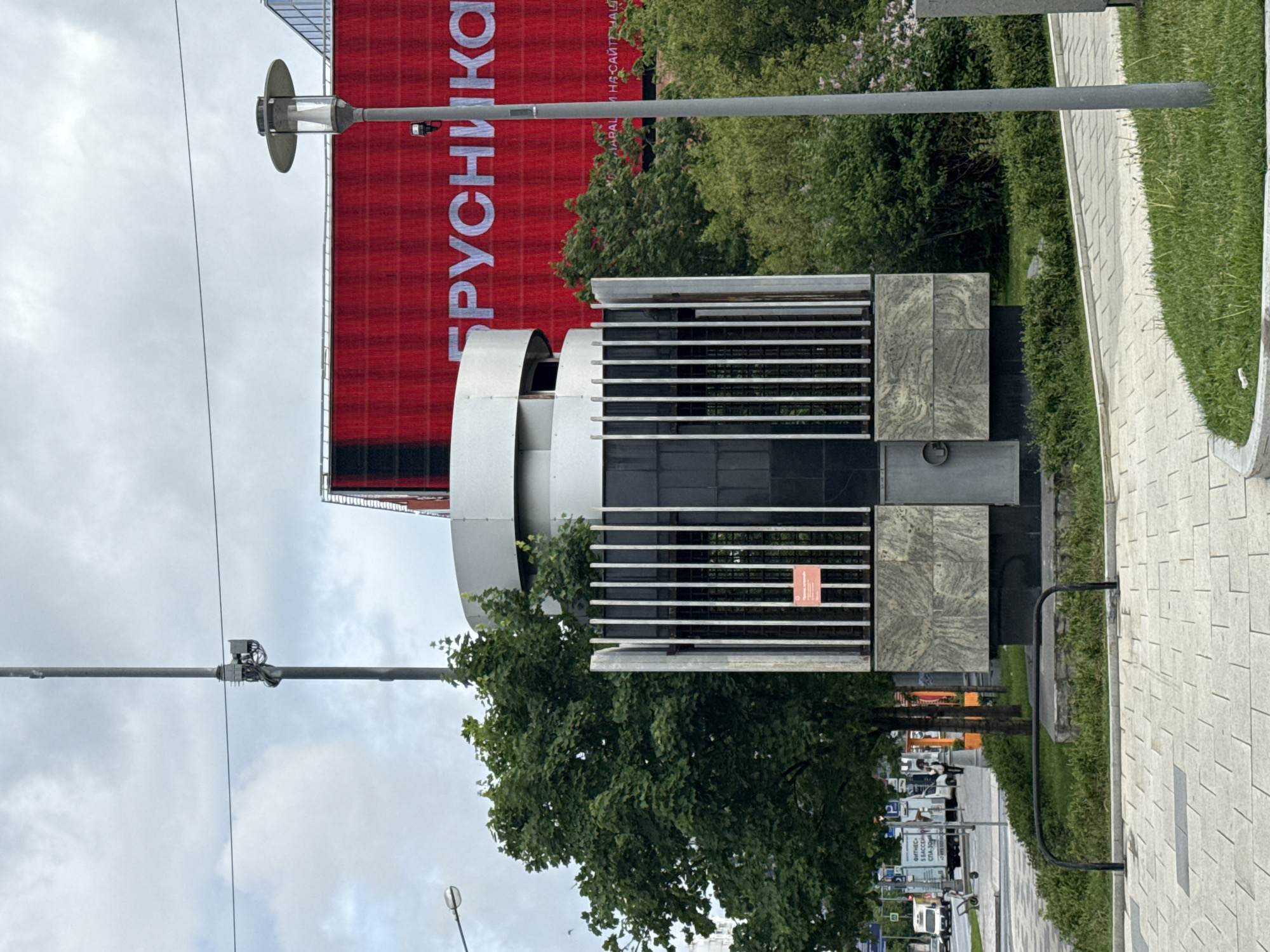 The image shows a street scene in Moscow, Russia. In the foreground is a modern sculpture consisting of white curved elements, black panels, and grey stone blocks with a horizontal slatted design. A small red sign is attached to the slatted section. Behind the sculpture is a large red billboard with the word "БРУСНИКА" in white Cyrillic letters. Trees partially obscure the view of the billboard. A street with light-colored pavement runs along the right side of the image, with a narrow strip of grass separating the pavement from the sculpture. In the background, cars, trucks, and buildings are visible along the street. A row of streetlights extends into the distance. The sky is overcast.