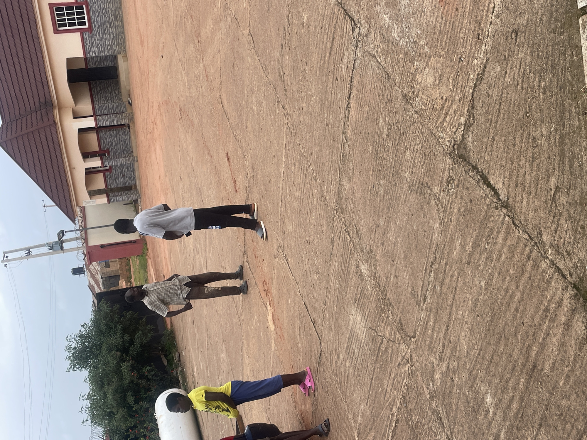 The image depicts a group of young men standing on a paved surface, likely an outdoor area or compound, with a building visible in the background. The building appears to be a residential structure with a tiled roof and windows. The ground is concrete with visible cracks and textures.
There are at least four individuals visible, all appearing to be young men or boys. One person in the foreground is wearing a yellow t-shirt and blue shorts, with pink flip-flops. Another person, further back, is wearing a checkered shirt and shorts. Two more individuals are further up the frame, one in a white t-shirt and black pants, and another in a white t-shirt. Their stances suggest they are standing around or perhaps engaged in a brief pause.
The scene is outdoors, and the lighting suggests it is daytime. The sky is overcast, indicating potentially cloudy weather. The general atmosphere seems casual. There is a large green bush to the left of the frame, and some electrical poles and wires are visible in the distance, suggesting an urban or semi-urban setting.
No specific activity or event is clearly identifiable beyond people standing around. The image is taken from a slightly elevated perspective, looking down at the subjects. There is no visible text within the frame. The context provided indicates the location is Jos, Nigeria.