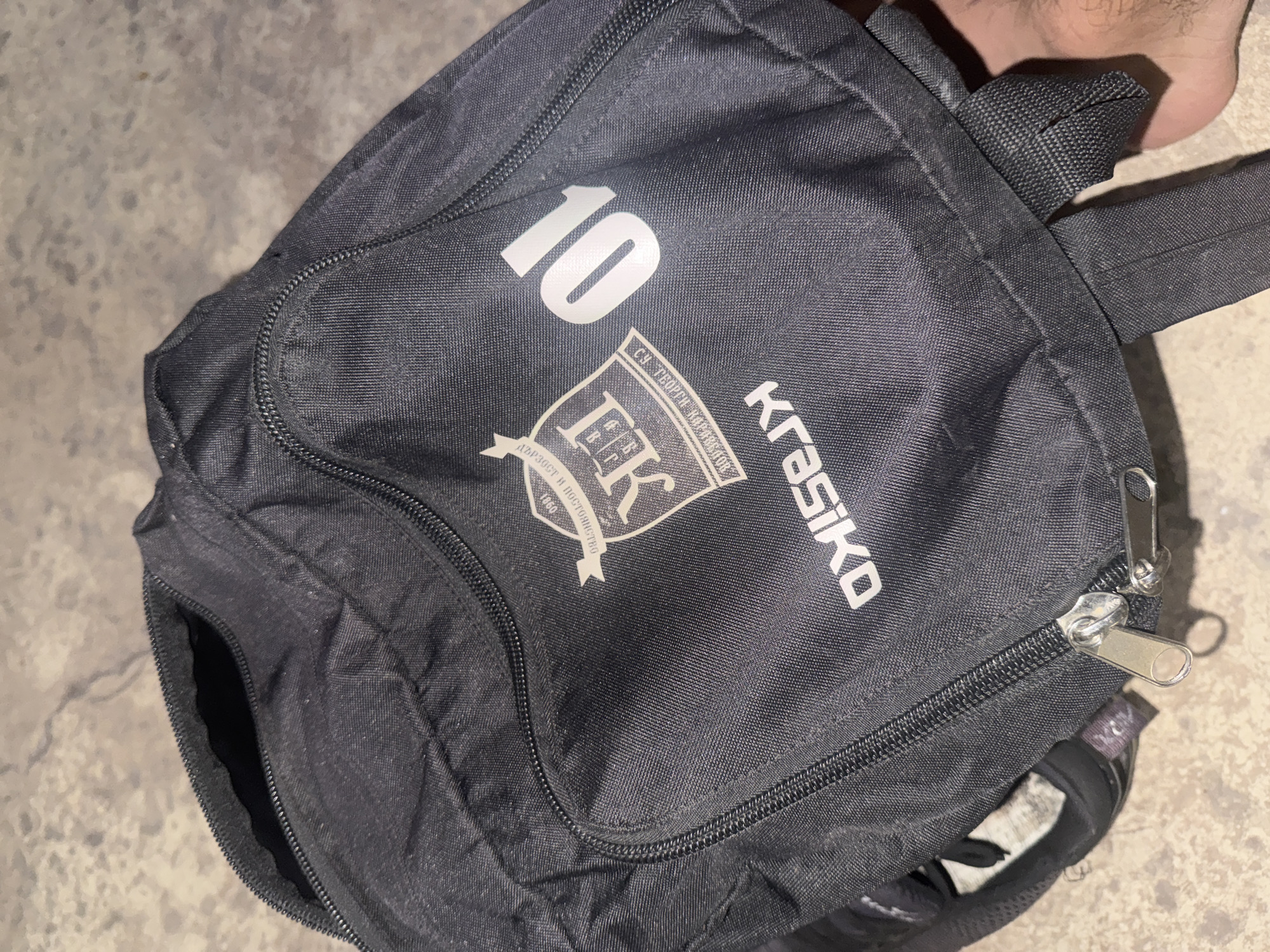 The image shows a black backpack with a white logo and the number "10" on its front pocket. The logo is a crest with the letters "FK" inside and text around it. Below the crest, the word "KRASIKO" is visible. The backpack is lying on a textured, concrete-like surface. A human foot is visible in the lower right corner of the image. The scene appears to be outdoors.