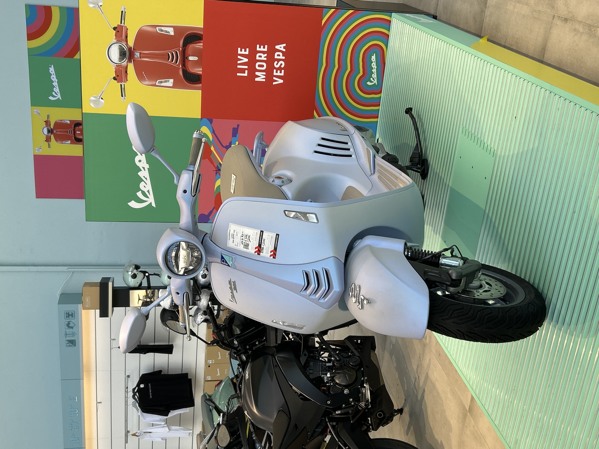 The image shows a light blue Vespa scooter prominently displayed in what appears to be a dealership or showroom. Behind the scooter, there's a vibrant display with Vespa-related graphics and the slogan "LIVE MORE VESPA." The scooters is resting on a striped green and yellow platform. To the left, a section of the store displays clothing, helmets, and other items related to Vespa, along with an indication of geographic coordinates: "10° 37' 10" E." Further in the background, partially obscured by the Vespa, is a black motorcycle. Overall, the scene captures a curated promotional setup for the Vespa brand within a retail environment.