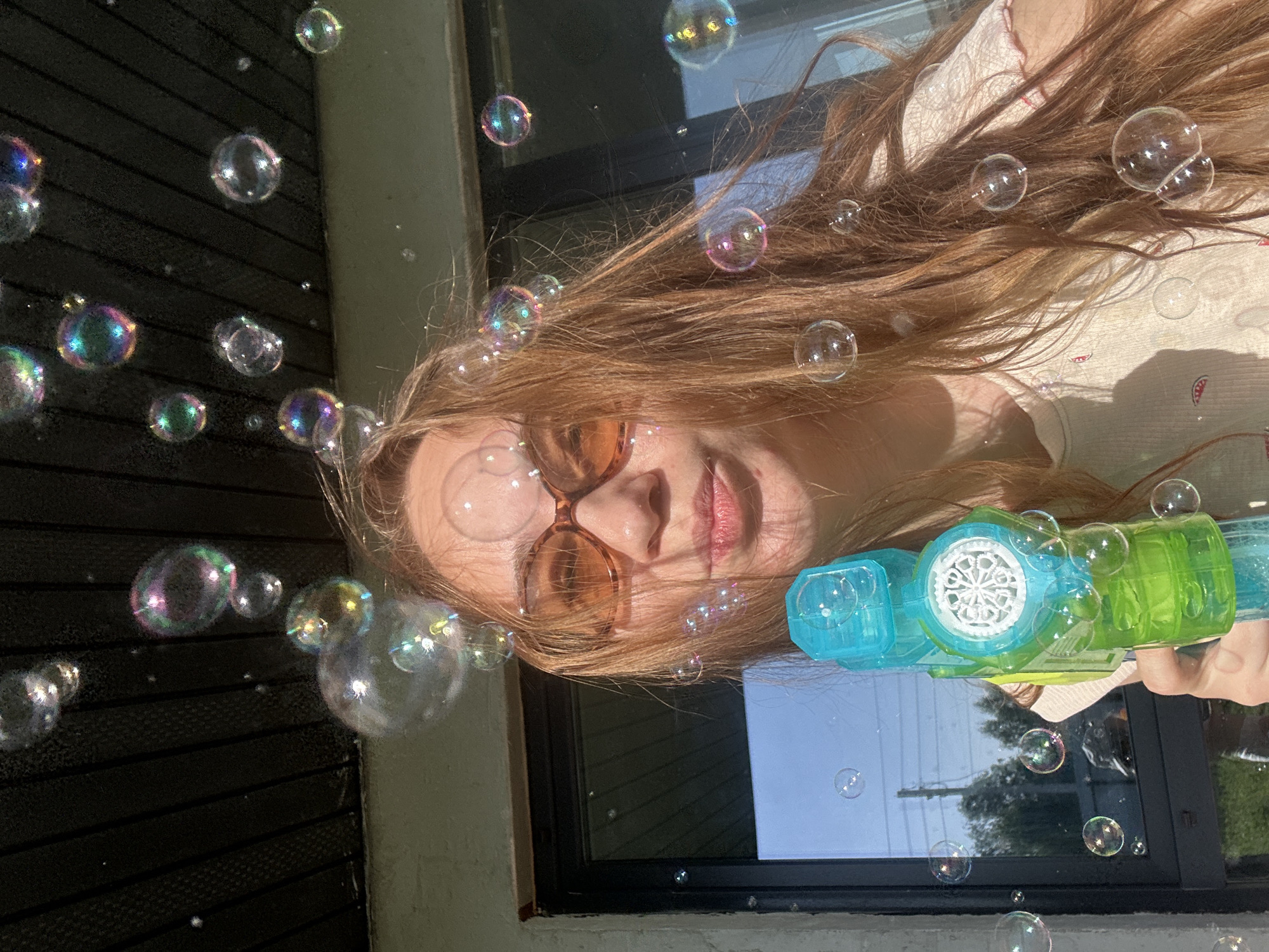 A person with reddish-brown hair, wearing brown sunglasses, is partially visible in this outdoor photograph. They are holding a green and blue bubble gun, which is positioned near a window. Several bubbles float in the air, some clustered to the left of the person's face and others dispersed around the bubble gun and window. The background consists of a dark, possibly slatted wall to the left and a window frame with a view of the sky and greenery visible through the glass. The scene appears to be taken during daylight. The location is unknown.