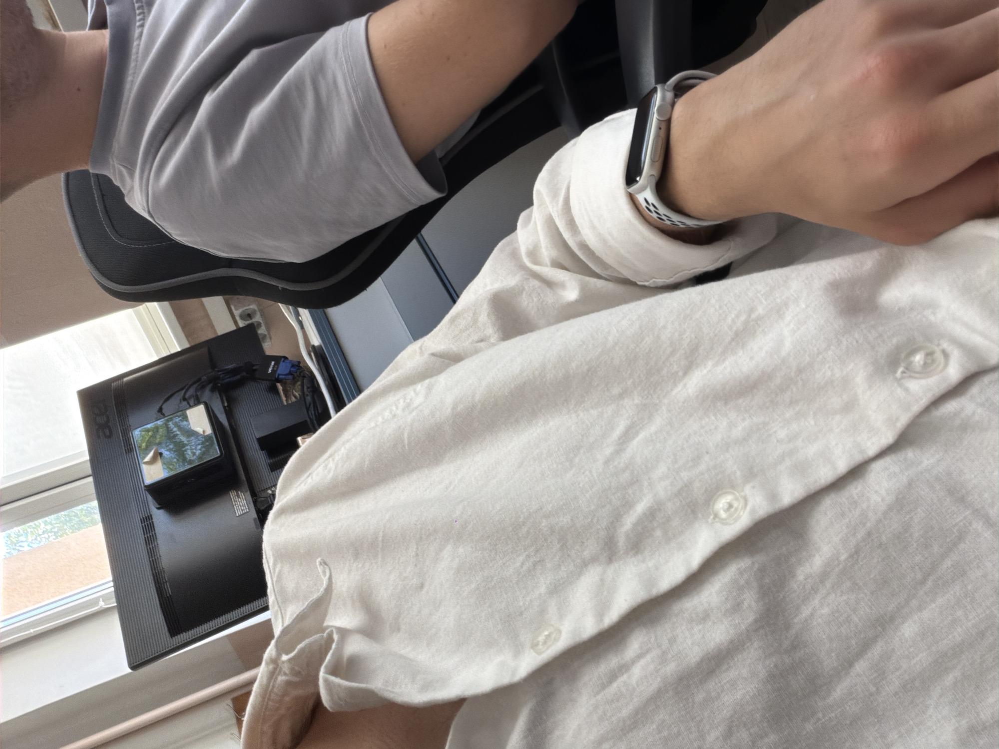The image shows a close-up of two individuals sitting at desks, likely in an office environment. The person in the foreground is wearing a white linen shirt and an Apple Watch on their left wrist. The person in the background is wearing a grey t-shirt. A computer monitor with the Acer logo is visible, along with what appears to be a small black box attached to the back of the monitor with tape. The scene is well-lit, suggesting it is daytime. There is no information in the image to suggest it is located in Plovdiv, Bulgaria; it is a generic office interior.