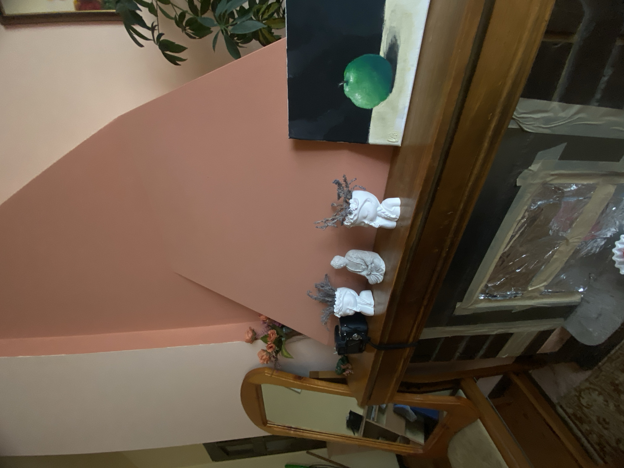 The image shows a corner of a room with light pink walls and a wooden shelf. On the shelf are several decorative items: a painting of a green apple, three small white sculptures, a vase with flowers, and a camera. There is also a mirror with a wooden frame below the shelf. The furniture is placed against a wall and there is a plant in the upper left corner. The location is not discernible from the image.