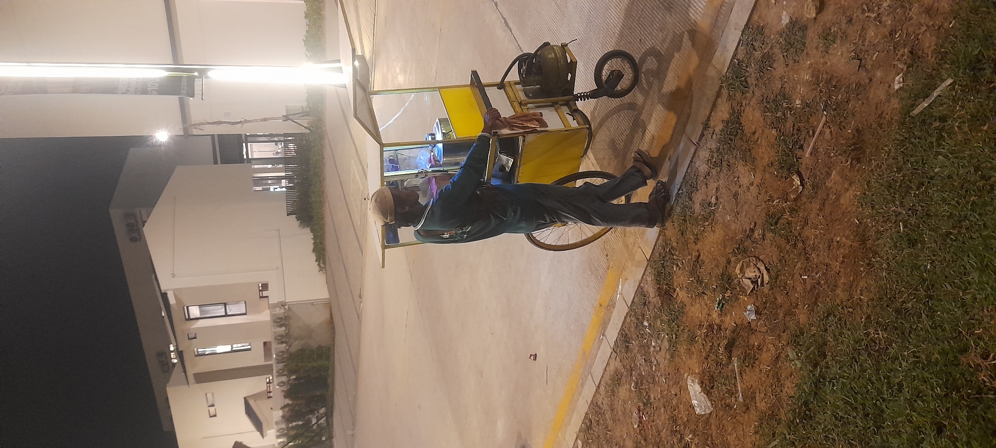 A man wearing a dark green long-sleeved shirt and dark pants with sandals is standing next to a food cart on a paved surface in Kragilan, Indonesia. The food cart is yellow with a clear plastic enclosure and has a canopy. The man is holding a cloth in his right hand. The cart has a large wheel on the left side and a smaller wheel with a spring suspension on the right. A gas tank is attached to the right side of the cart. The background shows modern-looking buildings with white walls and dark windows. There is a yellow line painted on the paved surface separating it from a dirt and grass area. The scene is lit by artificial lights, suggesting it is nighttime. Trash is visible on the ground in the dirt and grass area.