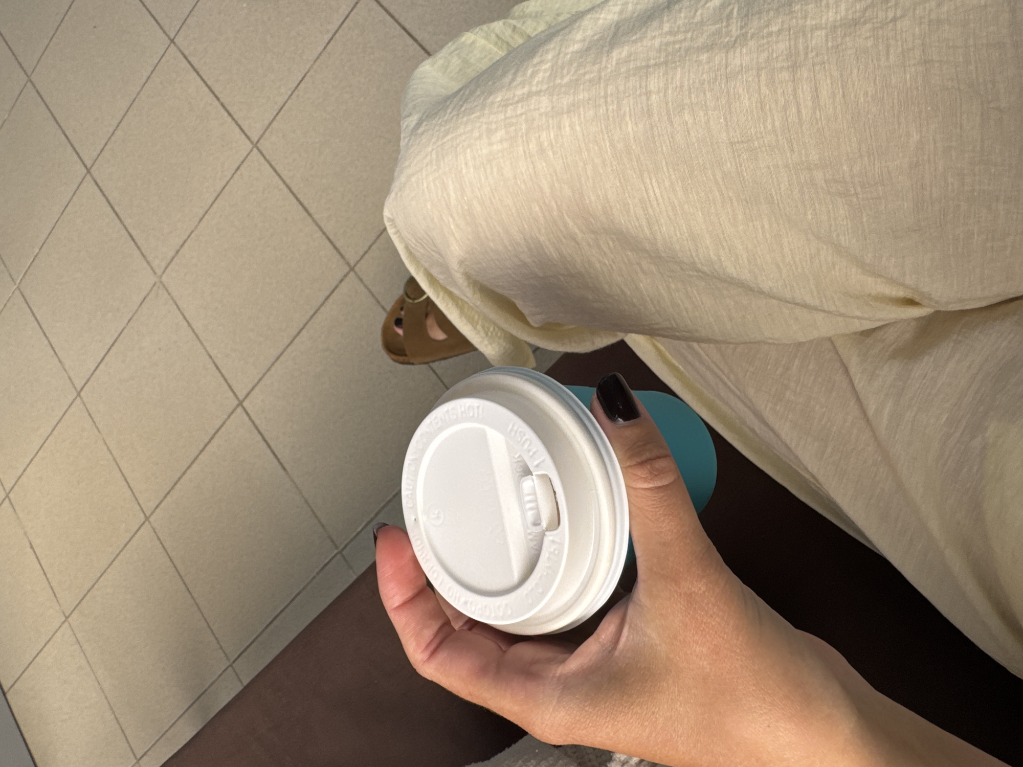A person's hand holds a turquoise coffee cup with a white lid. The lid has "CAUTION CONTENTS HOT" and "PULL TO OPEN" printed on it. The person's nails are painted black. They are wearing a light-colored, loose-fitting garment and brown sandals. The background consists of tiled flooring and a dark surface, possibly a bench. The scene is indoors, likely in a public space like a cafe or waiting area. The image does not provide information about the city of Chaikovsky, Russia.