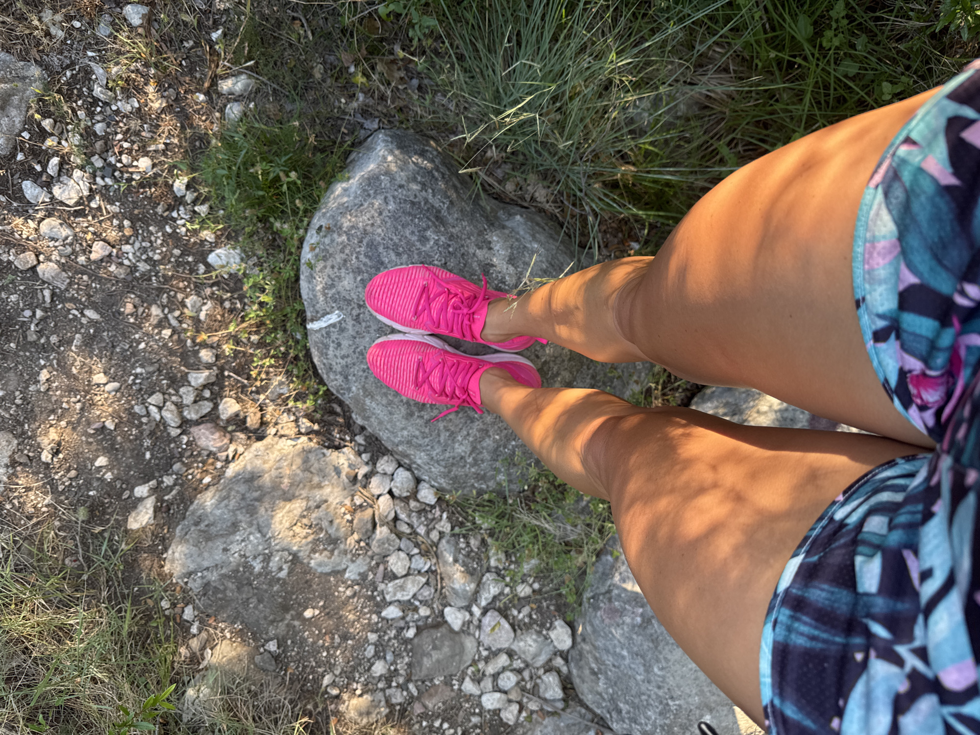 A pair of legs wearing pink running shoes and patterned shorts are positioned on a rocky trail. The legs are tanned and illuminated by dappled sunlight, casting shadows on the skin. The surrounding environment consists of dry ground, scattered rocks, and patches of green grass. The scene appears to be outdoors in a natural setting, potentially a hiking or running path. There is no specific indication of the city of Rafailovići, Montenegro.