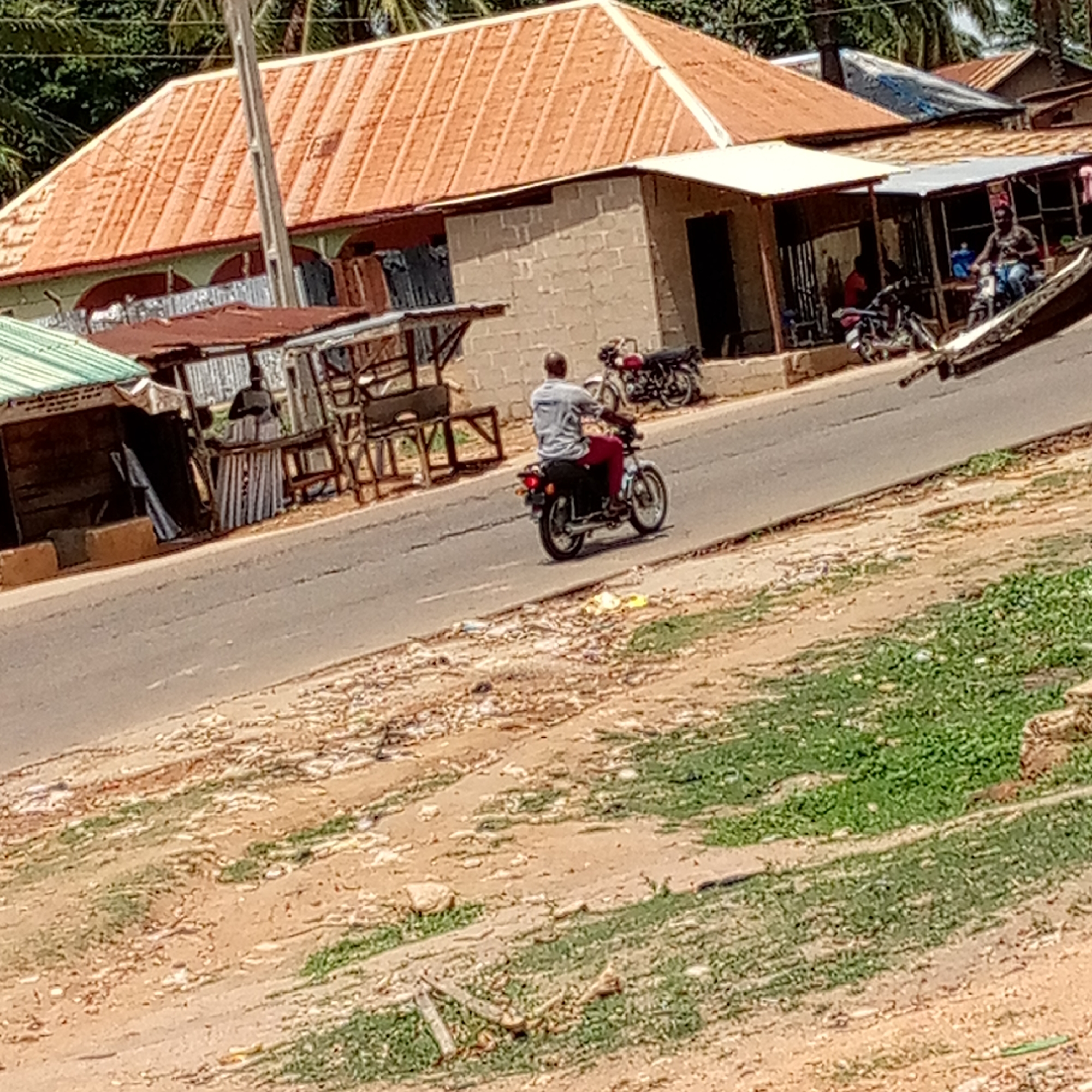 The image captures a daytime scene along a street in what appears to be a rural or semi-urban area. In the center, a man is seen from behind, riding a motorcycle down a paved, slightly sloping road. He wears a light-colored shirt and red pants. Another black and red motorcycle is parked on the right side of the road, in front of a building.
The background features several structures. On the left, there are small, simple stalls or shops with green and rusted corrugated metal roofs, and wooden frames. One stall appears to have a dark figure or mannequin behind a counter made of corrugated sheets. To the right, a more substantial building with a prominent orange-tiled roof and light-colored walls stands, alongside other smaller structures with various types of roofs, some also corrugated metal. Palm trees are visible in the distance, suggesting a warm climate. A utility pole stands tall towards the upper center.
The foreground consists of a dirt and gravel shoulder beside the road, with patches of green vegetation and small rocks. The lighting is bright, indicating a sunny day, and distinct shadows are visible, suggesting mid-morning or afternoon. There is no discernible text visible in the image.