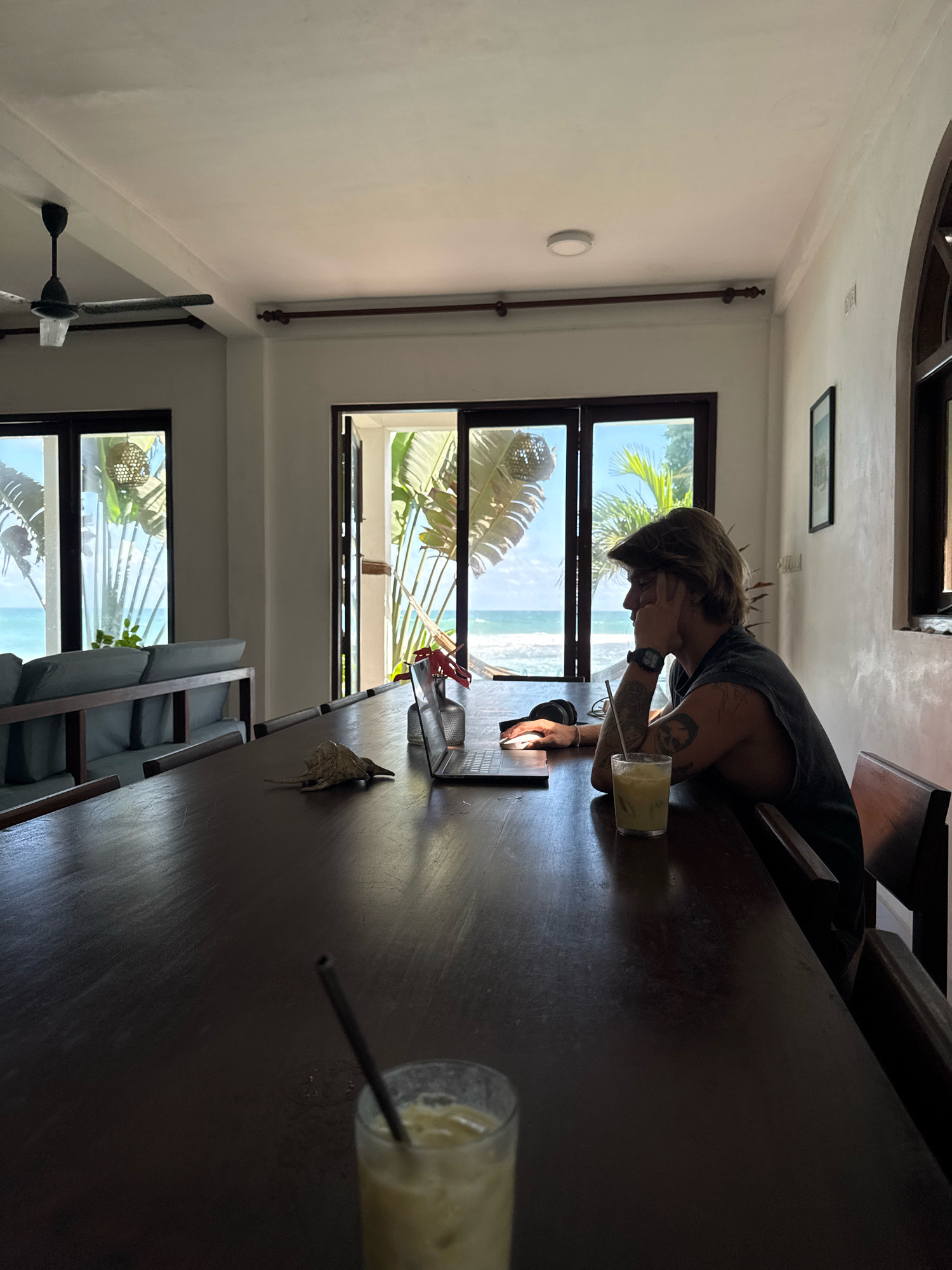 A young man with shoulder-length blonde hair, wearing a dark, sleeveless t-shirt, sits at a dark wooden table in what appears to be a room with large windows looking out onto a tropical beach. He is facing to the right, with his left arm resting on his chin, and his right arm on the table. He is wearing a watch on his left wrist and has tattoos on his right arm. A laptop is open in front of him on the table, along with a glass of light-colored liquid with a straw, a metal vase with a few red flowers, and a decorative shell. Another glass of the same beverage with a straw is visible in the foreground, out of focus. In the background, through the windows, palm trees and the ocean are visible. A ceiling fan is mounted on the ceiling. The room has white walls and a wooden ceiling.