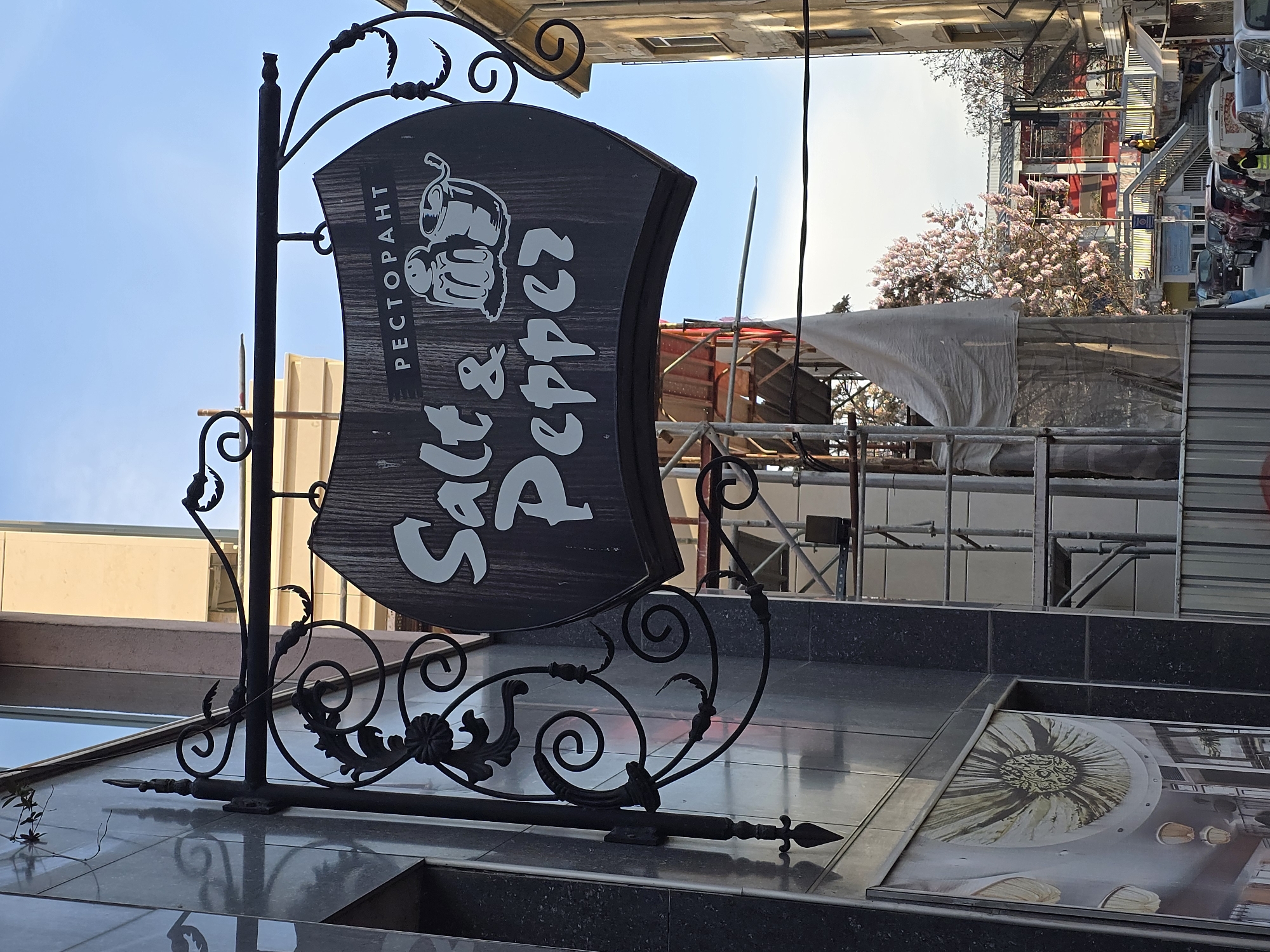 The image depicts a sign for "Salt & Pepper" restaurant, written in white letters on a dark, irregularly shaped board framed by ornate black metalwork. The word "PECTOPAHT" is written vertically in Cyrillic on the upper left side of the board. The sign is likely mounted on the facade of a building, with a reflective surface below showing a partial image of what appears to be an advertisement or design. In the background, there is construction work with scaffolding and a protective white tarp. Beyond the construction, a building with a red facade is visible, along with a blossoming tree. The sky is pale blue, suggesting daylight. The image appears to be taken in an urban setting.