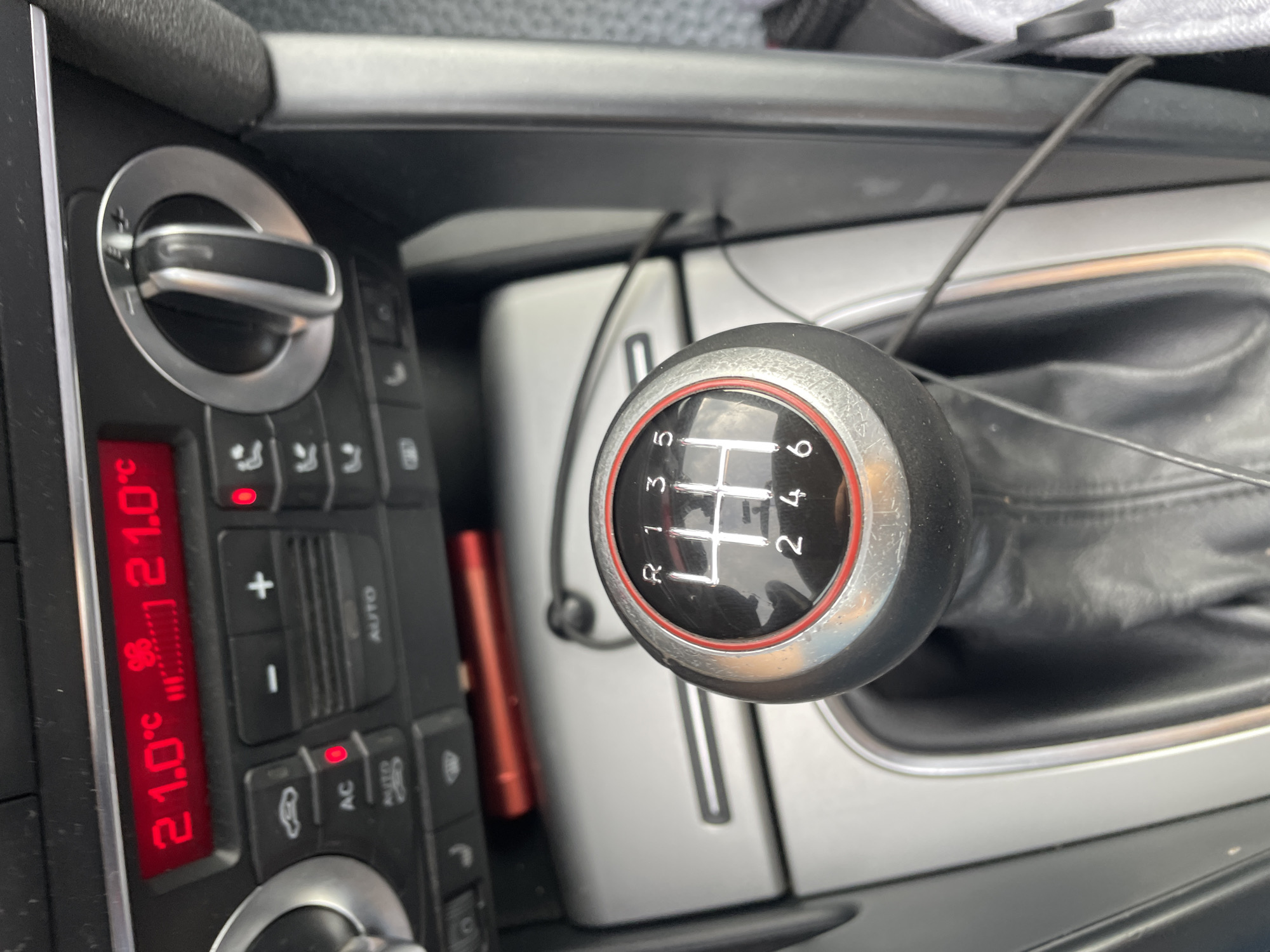 This is a close-up, slightly angled shot of a car's interior, focusing on the center console. The primary subjects are a manual transmission gear shifter and climate control buttons. The gear shifter, a black knob with a chrome and red trim, displays a six-speed manual gear pattern (1, 2, 3, 4, 5, 6, and R for reverse). It appears to be in neutral or a similar position.
To the left of the gear shifter are the climate control elements. A digital display shows "21.0°C" in red, indicating the temperature. Below this are several buttons with icons and text, including controls for temperature adjustment (+/-), fan speed, air conditioning (AC), and auto mode. A silver dial with a black grip is also visible, likely for temperature control.
The background shows the textured fabric of the car's upholstery and a portion of the dashboard. A black cable is visible running across the console, possibly for charging a device or connecting to the infotainment system. The overall scene suggests the interior of a modern passenger car.
There are no people visible in the image. The lighting suggests it might be daytime, but without external views, the weather and precise time of day are indeterminate. The location context provided is Criuleni, Moldova, which, while not directly evident from visual cues within the image, provides a geographical setting. No strong emotions are conveyed; the image is factual and observational. Visible text includes "AUTO", "AC", and the temperature reading "21.0°C". The condition of the gear shifter suggests it has been used, with some minor scuffing visible on the chrome ring.