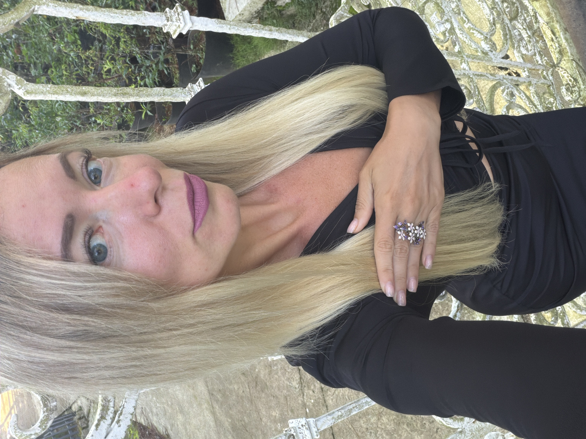 A woman with blonde hair and blue eyes, wearing a black dress and a ring on her finger, is taking a selfie in what appears to be a garden setting. The background consists of greenery and some white ornate structures, possibly a fence or railings. The location is specified as Xuân Trường - Đà Lạt, Vietnam.