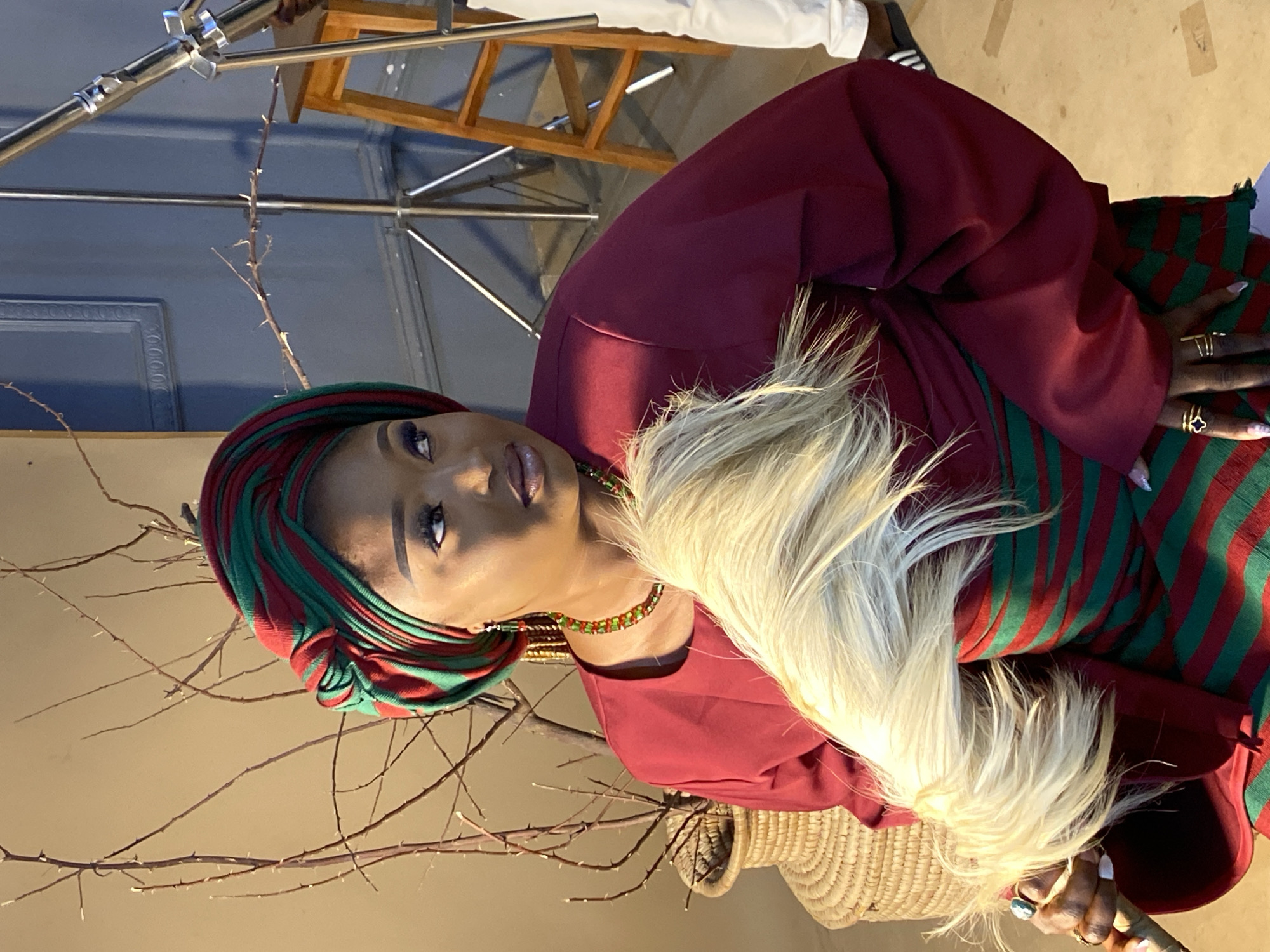 The image shows a woman in traditional attire, specifically a red and green striped headwrap and dress, holding a whisk made of light-colored hair. She is wearing a beaded necklace and earrings. The background appears to be a studio setting with a blue textured wall, a wooden stool, and photography equipment, including a light stand. A woven basket and dried branches are also visible. The woman is looking to her right, with her makeup clearly visible. The scene is lit by studio lights, casting shadows. The location is identified as Bukuru, Nigeria.