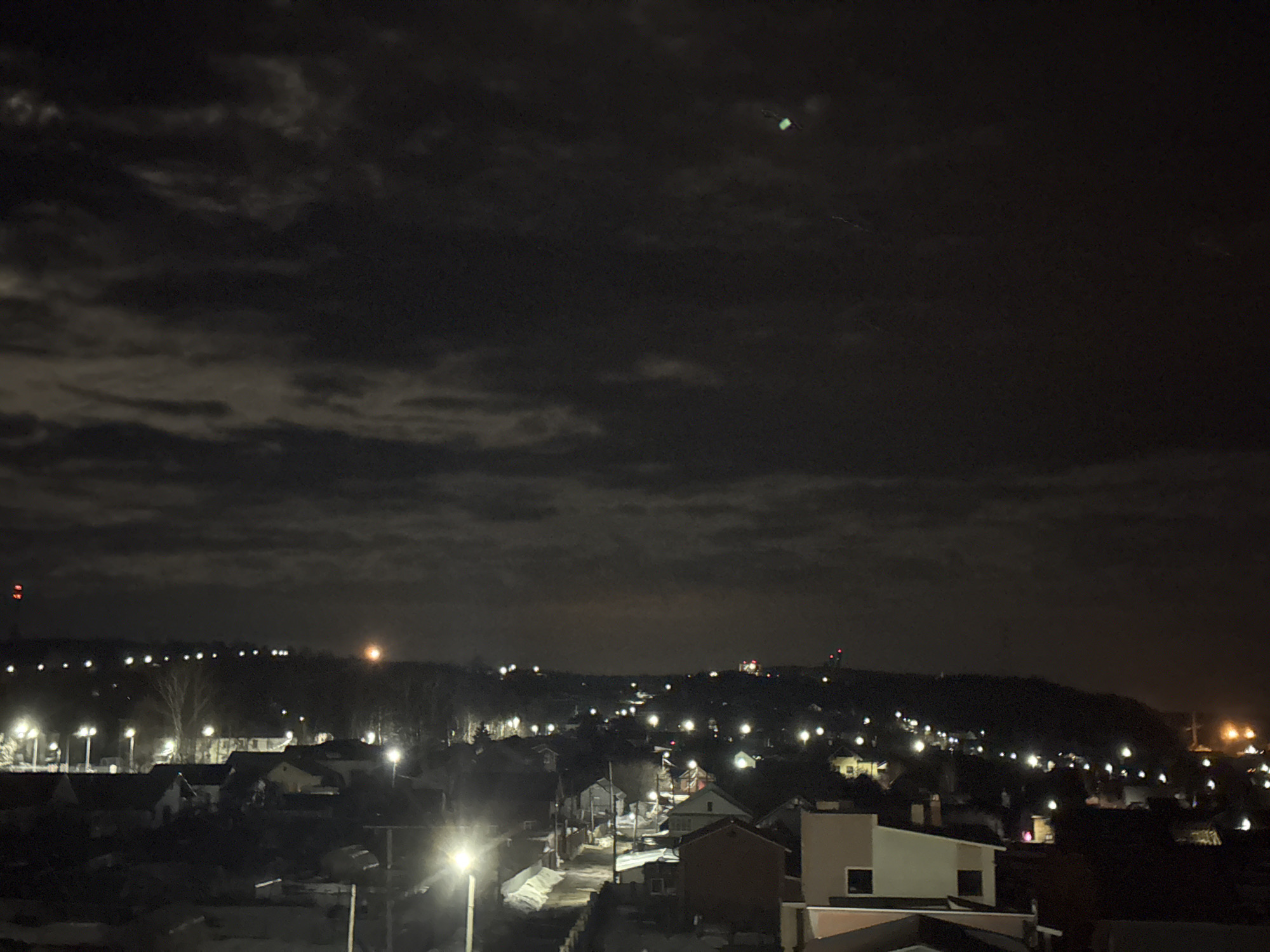 The image depicts a night scene in the city of Chaikovsky, Russia. The sky is dark and cloudy, with a dim green light visible in the upper portion of the frame. Below the sky, a townscape is visible, punctuated by numerous lights. A series of lights line the horizon, suggesting streetlights or the lights of buildings on a distant hill. In the foreground, streets illuminated by streetlights lead through a residential area. Buildings of varying sizes and shapes are scattered throughout the scene. Snow covers some of the ground and rooftops. The image evokes a quiet, suburban environment at night.