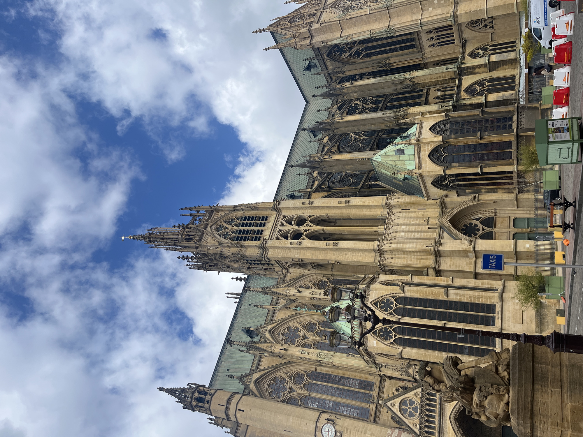 This is a ground-level, upward-angled, color photograph of the Metz Cathedral, also known as the Cathedral of Saint Stephen of Metz, in Metz, France. The cathedral is the main subject of the image, occupying the majority of the frame. Its beige stone facade is visible with intricate gothic architectural details, including arched windows, buttresses, spires, and sculpted elements. The roof is covered in green patina. The blue sky speckled with white clouds forms the backdrop on the left side.
On the right side of the image, a section of the street and adjacent sidewalk is visible. There is a blue TAXIS sign on a pole, a light post, and a partially visible sculpture. In the background, there is a row of red and white barriers along the street and a white van with the company name "APIB" in blue. There appears to be a person in dark clothing working on the pavement.