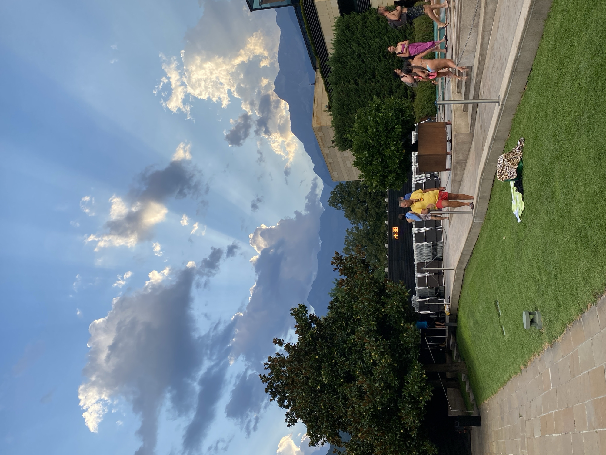 The image depicts a scene at a public swimming pool complex, likely in Meran - Merano, Italy, given the presence of mountainous terrain and the overall aesthetic. The sky is a vibrant blue with dramatic sunbeams piercing through scattered clouds, creating a picturesque natural light. In the foreground, a well-maintained lawn with a paved pathway transitions to a tiered concrete area around the pool.
Several individuals are visible, some in swimwear, indicating they are using the pool facilities. A man in a yellow shirt and red shorts stands near the pathway, appearing to be an employee or lifeguard. A group of people, including women in bikinis and one in a pink dress, are walking up a set of stairs. In the background, there are lounge chairs, a dark-colored building with an illuminated digital display showing "rh 38", and lush green trees. The backdrop features a range of imposing mountains, shrouded in a light haze, characteristic of the alpine region. The overall atmosphere suggests a relaxed, recreational setting during daylight hours, possibly late afternoon due to the quality of light.
