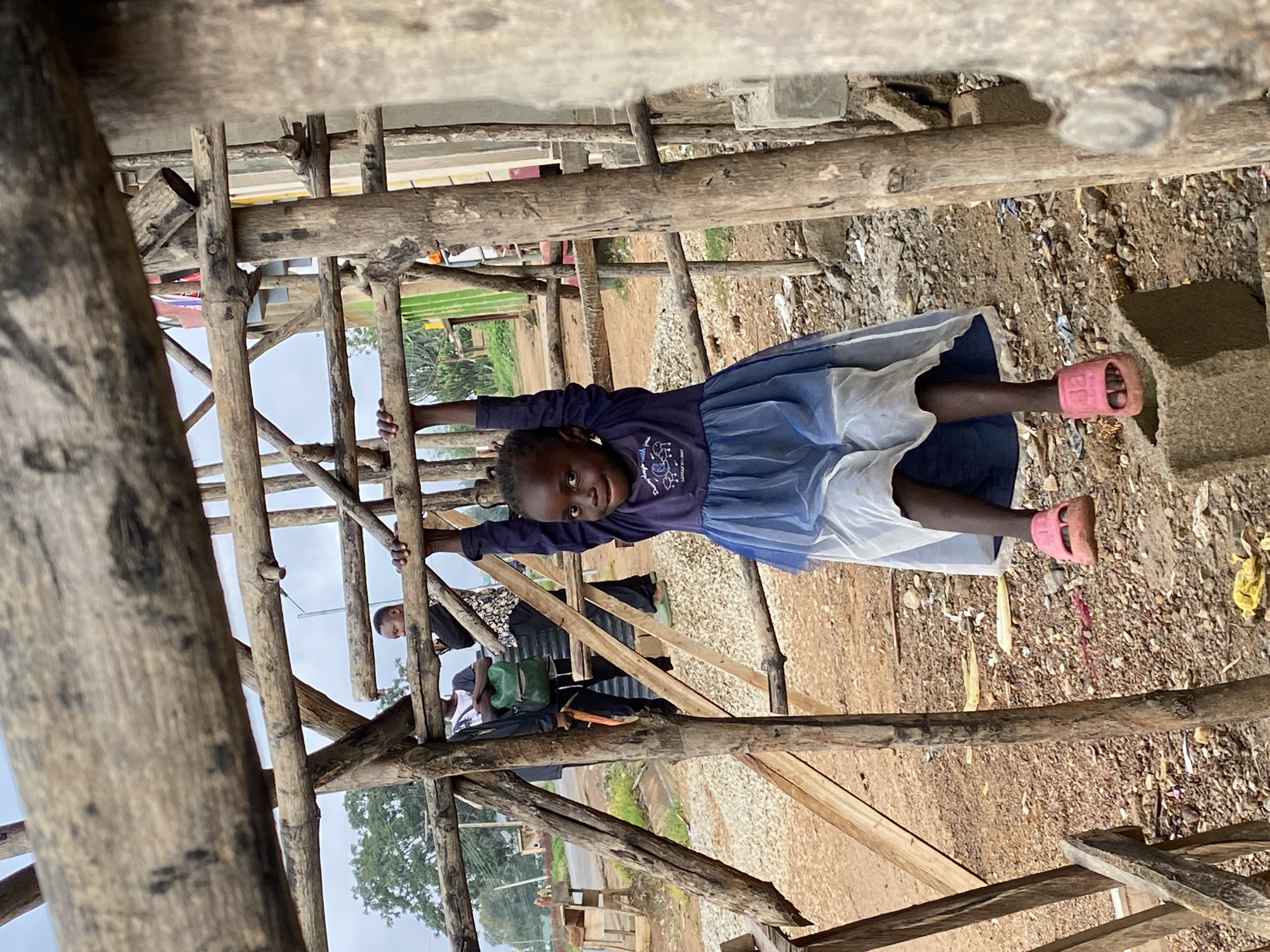 The image, rotated 90 degrees clockwise, captures a cheerful young Black child holding onto a wooden structure in Riyom, Nigeria. The child, positioned centrally, looks directly at the viewer with a broad, genuine smile, conveying a sense of joy or playfulness. She is dressed in a dark blue long-sleeved top that features white text, partially legible as "magic with" followed by "LOVELY LITTLE" below. She wears a light blue or white skirt/dress and bright pink open-toed sandals.
The child is seen suspended or leaning on rough-hewn wooden beams that form a scaffolding-like structure, filling much of the foreground and midground. Behind her and to the sides, other individuals are partially visible amidst the wooden framework; one adult's hand is seen gripping a beam, and another person is partially obscured by the structure.
The ground beneath the structure is a mix of dirt and gravel, with some small pieces of debris scattered around. In the background, beyond the immediate wooden framework, there are distant trees and what appear to be other buildings or landscape features under an overcast sky, indicating it is daytime. The setting suggests an outdoor environment, possibly a construction site or a temporary settlement in a developing area.