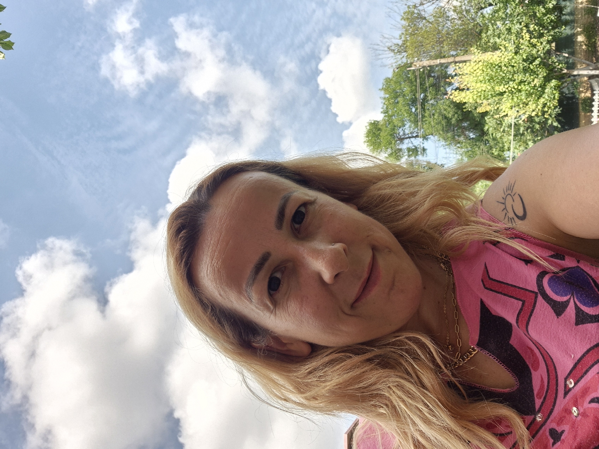A medium shot from a low angle shows a woman with blonde hair and light skin posing for a selfie. She is wearing a pink sleeveless top with a colorful graphic print and a gold chain necklace. A tattoo of a crescent moon and sun is visible on her left shoulder. The background consists of a blue sky with white clouds, and trees and a utility pole are visible on the right. The scene appears to be outdoors in a residential area.