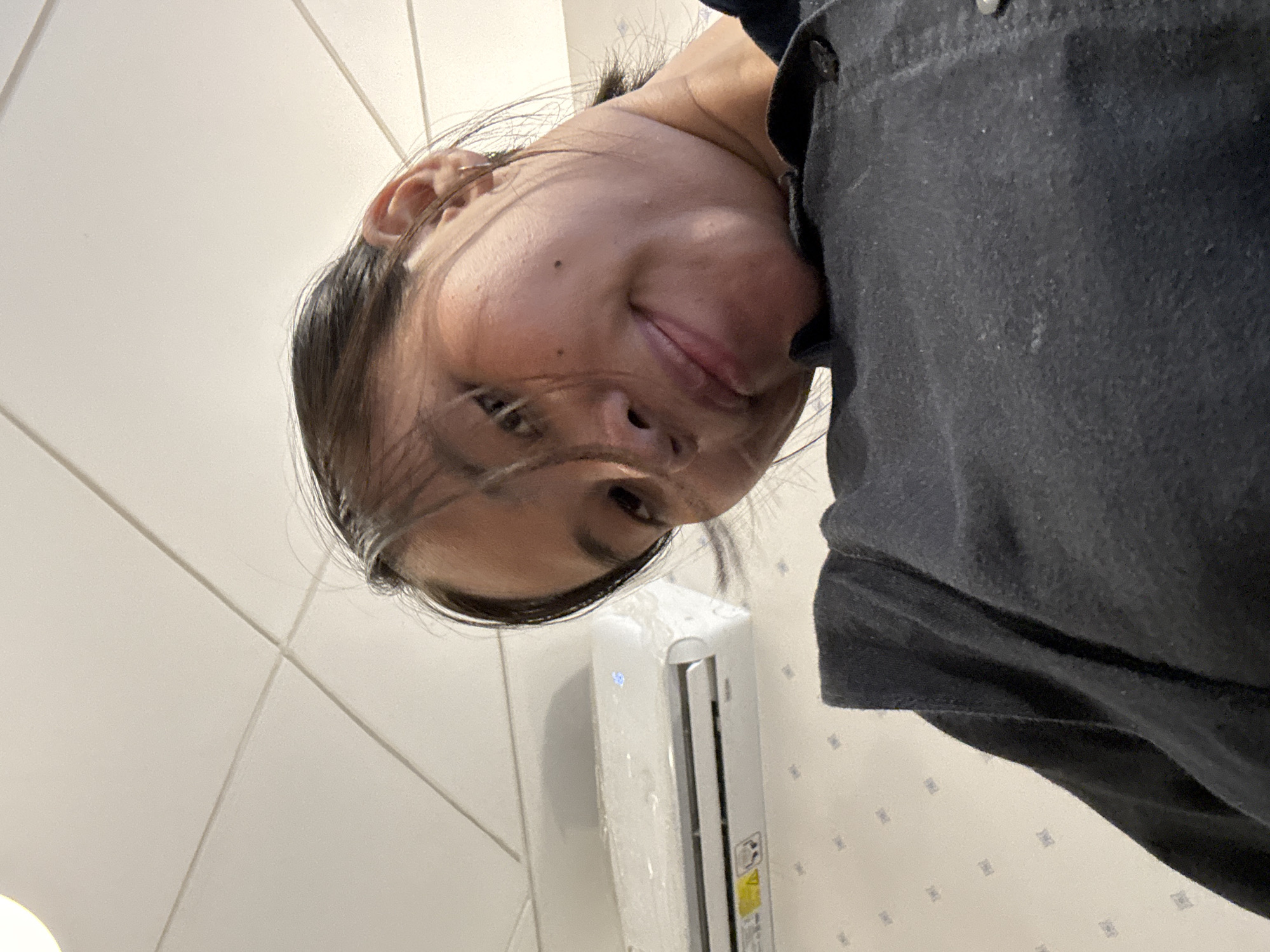 A low-angle, close-up, head-and-shoulders shot of a young Asian woman with dark hair, looking down at the camera. She is wearing a dark grey collared shirt. Behind her, a white air conditioning unit is mounted on a patterned wall, below a textured, tiled ceiling. The woman's face is tilted, with one eye partially visible. The lighting appears to be artificial and overhead. The location is unknown but the image metadata indicates it was taken in South Jakarta, Indonesia.