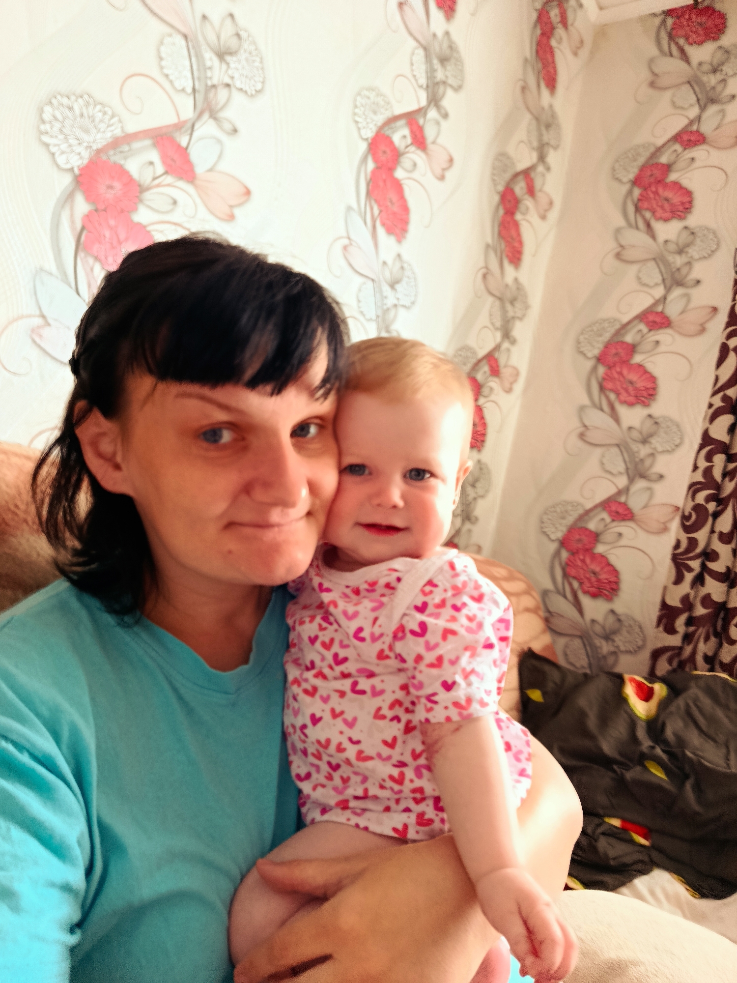This is a portrait photograph taken indoors, featuring a woman holding a baby. The woman, on the left, has dark hair with bangs, blue eyes, and a light complexion, wearing a light blue short-sleeved t-shirt. She is looking towards the camera with a slight smile.
The baby, held in the woman's arms, has light blonde hair, blue eyes, and a fair complexion. The baby is wearing a white short-sleeved top covered in small red and pink heart patterns, and is also looking at the camera with a gentle smile. Both individuals appear content and happy.
The background features patterned wallpaper with repeating floral motifs in shades of red, pink, and grey on a light-colored ground. To the right, a dark brown curtain with an intricate pattern is visible, along with a portion of a dark blanket or fabric item with some yellow and red shapes. The scene depicts a warm, intimate moment in what appears to be a home setting, likely during the daytime in Фоки, Russia. There is no visible text in the image.