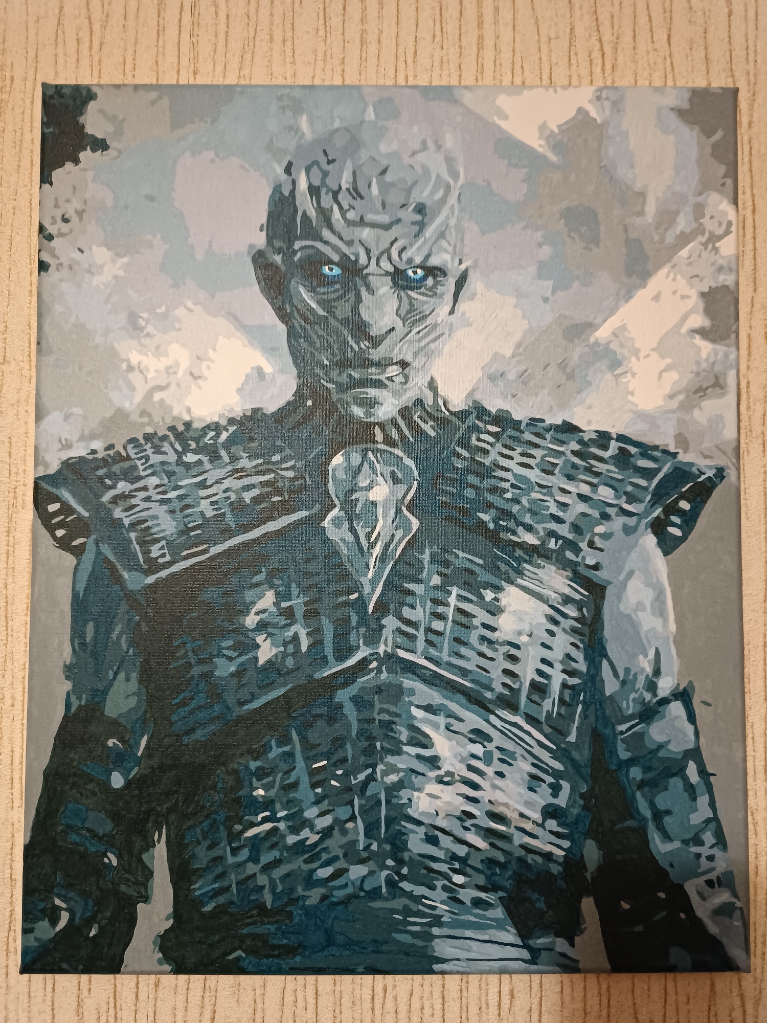 The image is a painting of the Night King from Game of Thrones. The painting is of a bust, showing the Night King from the shoulders up. He has pale, cracked skin, and his eyes are glowing blue. He is wearing armor that appears to be made of dark, scaled material, with a lighter, crystalline piece on his chest. The background is a muted gray and white, suggesting a cold, snowy environment. The painting is on canvas and is framed by a textured, beige material, possibly wallpaper. There is no indication of the city of Mohyliv-Podilskyi, Ukraine, in this image.